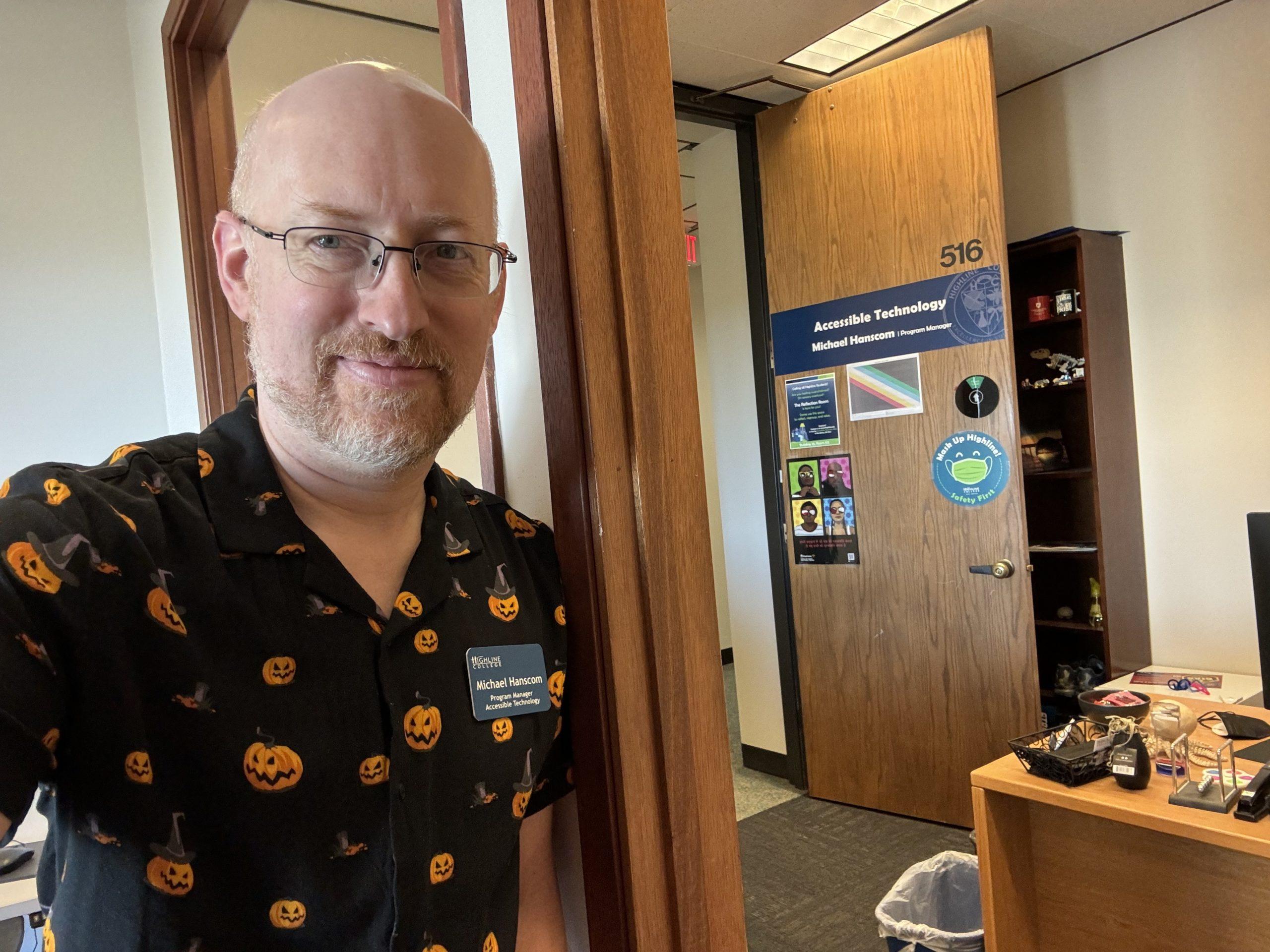 Me in my work office, wearing a black short-sleeve button-up shirt with orange jack-o-lanterns printed on it.