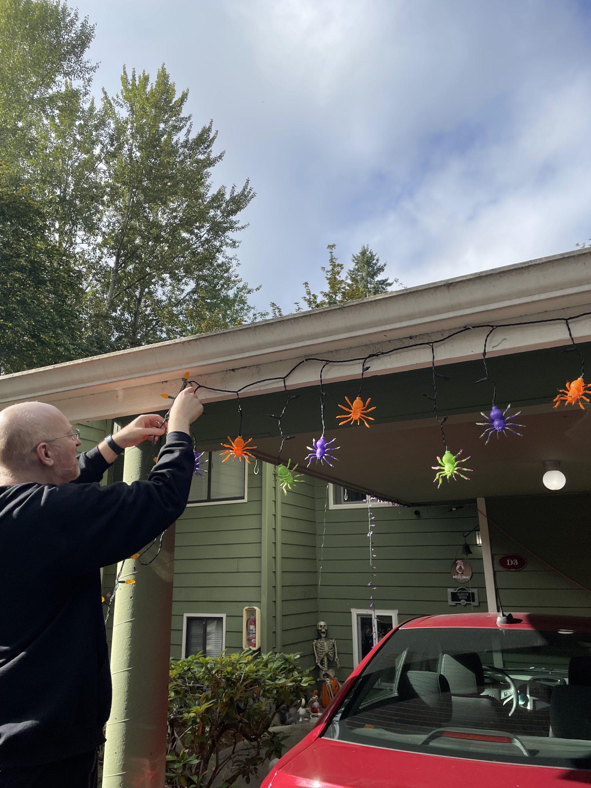 Me hanging Halloween lights with orange, green, and purple spiders from the carport in front of our house.