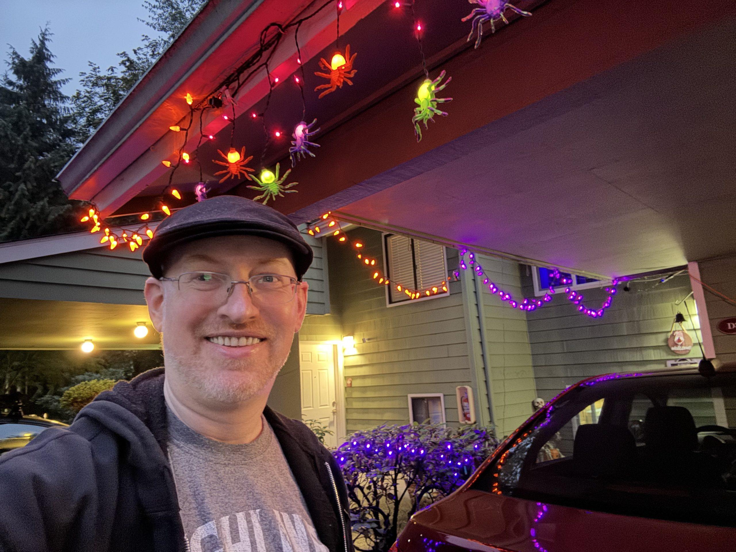 Me in front of our house with the halloween lights I put up yesterday glowing. There are purple and orange strings along one side, and glowing green, orange, and purple spiders along another.