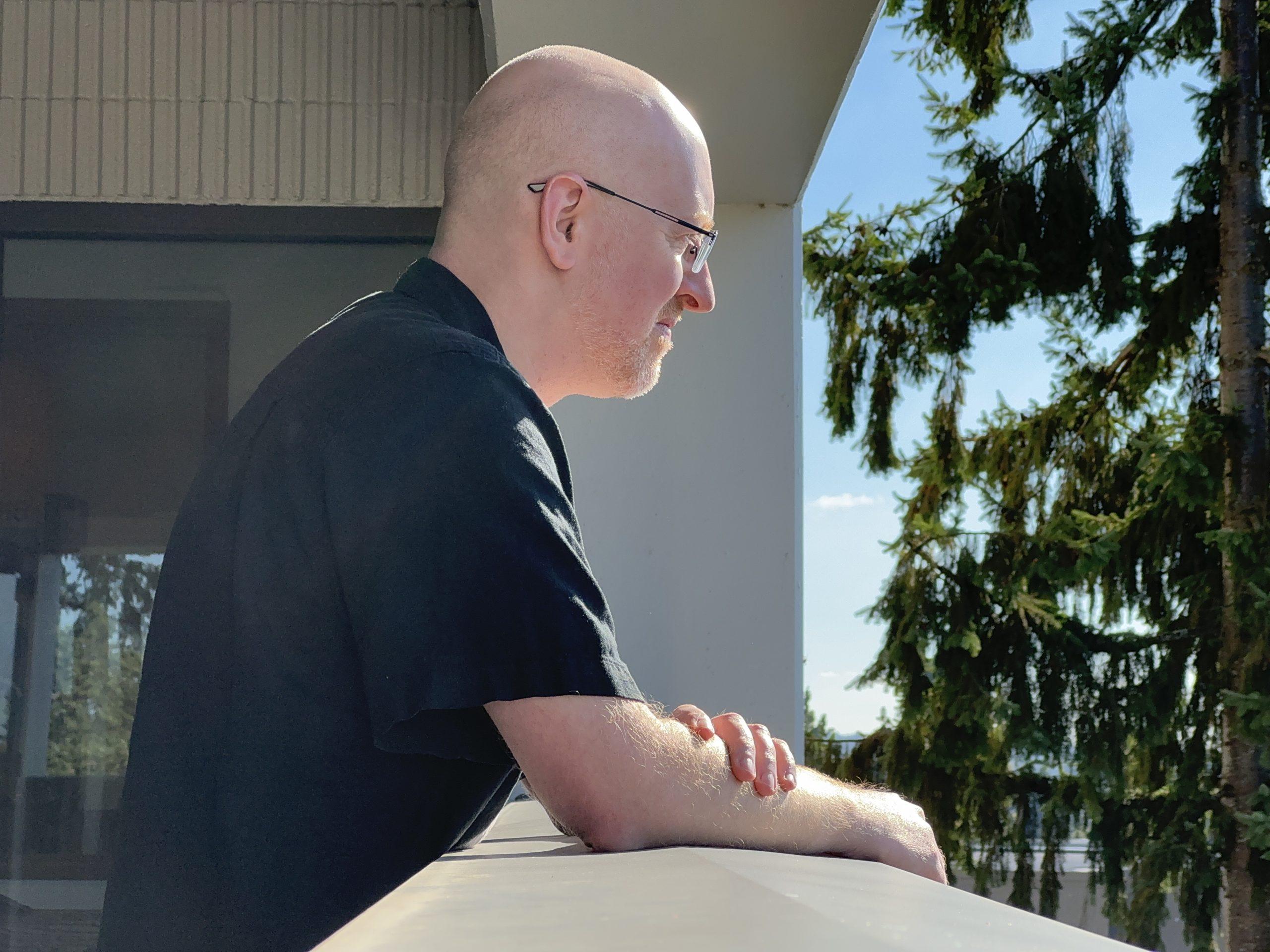 Me shot from my right side, leaning on a balcony railing and looking out away from the building.