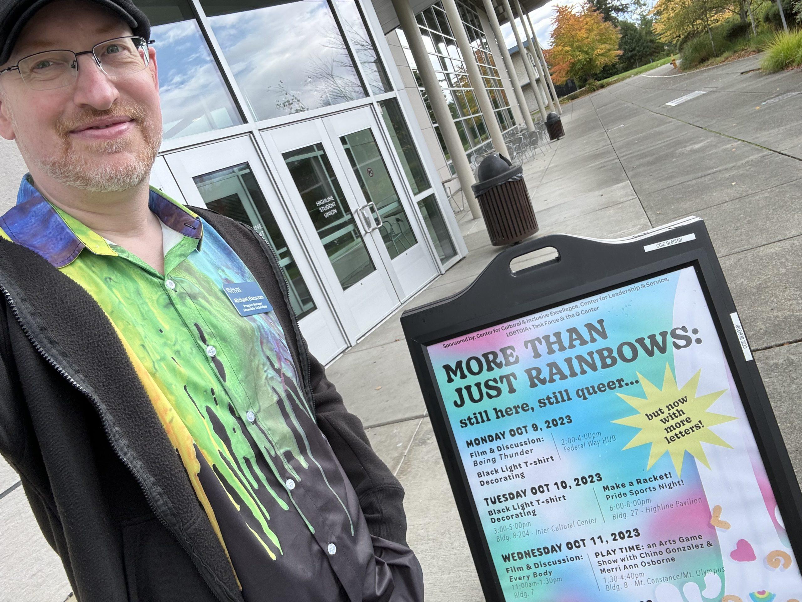 Me standing outside a building on the college campus, next to a sign listing events for LGBTQ week with the headline, 'More than just rainbows: still here, still queer, but now with more letters!'