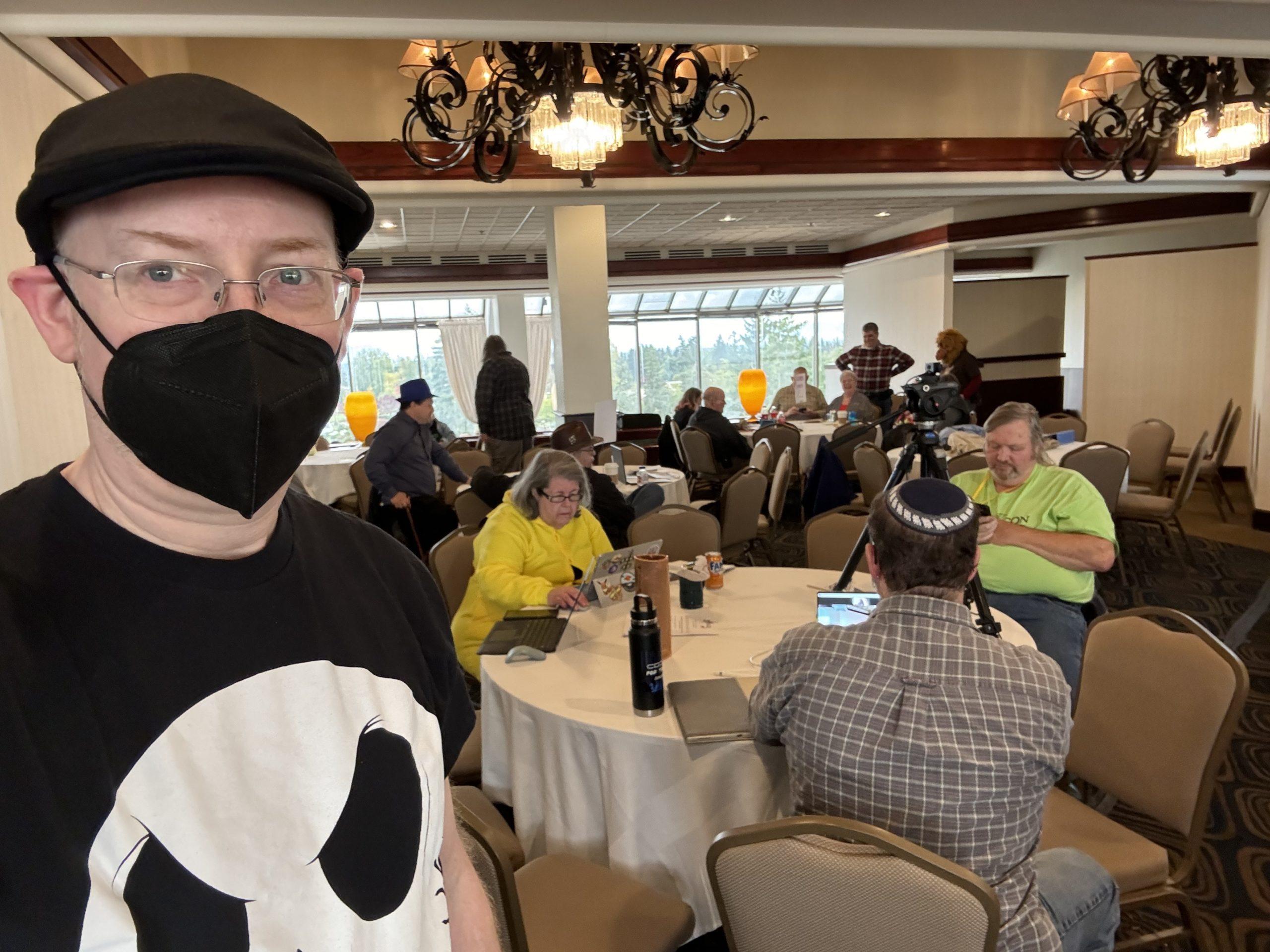 Me, wearing a Jack Skellington t-shirt and black facemask, standing in a hotel meeting room with a number of other people sitting at tables and chatting with each other behind me.