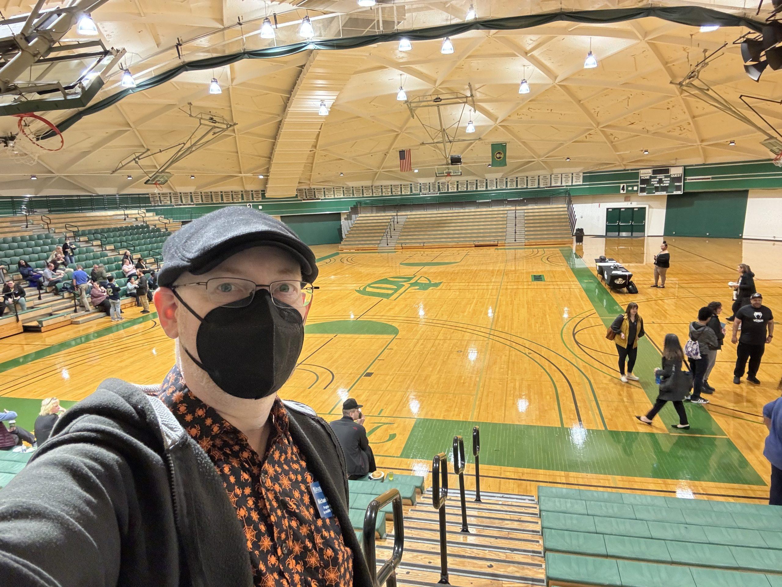 Me, wearing a orange-and black spider and spiderweb print shirt, black hoodie, black cap, and black mask, standing at the top of bleachers in a dome-roofed gym with the gym floor visible behind me.