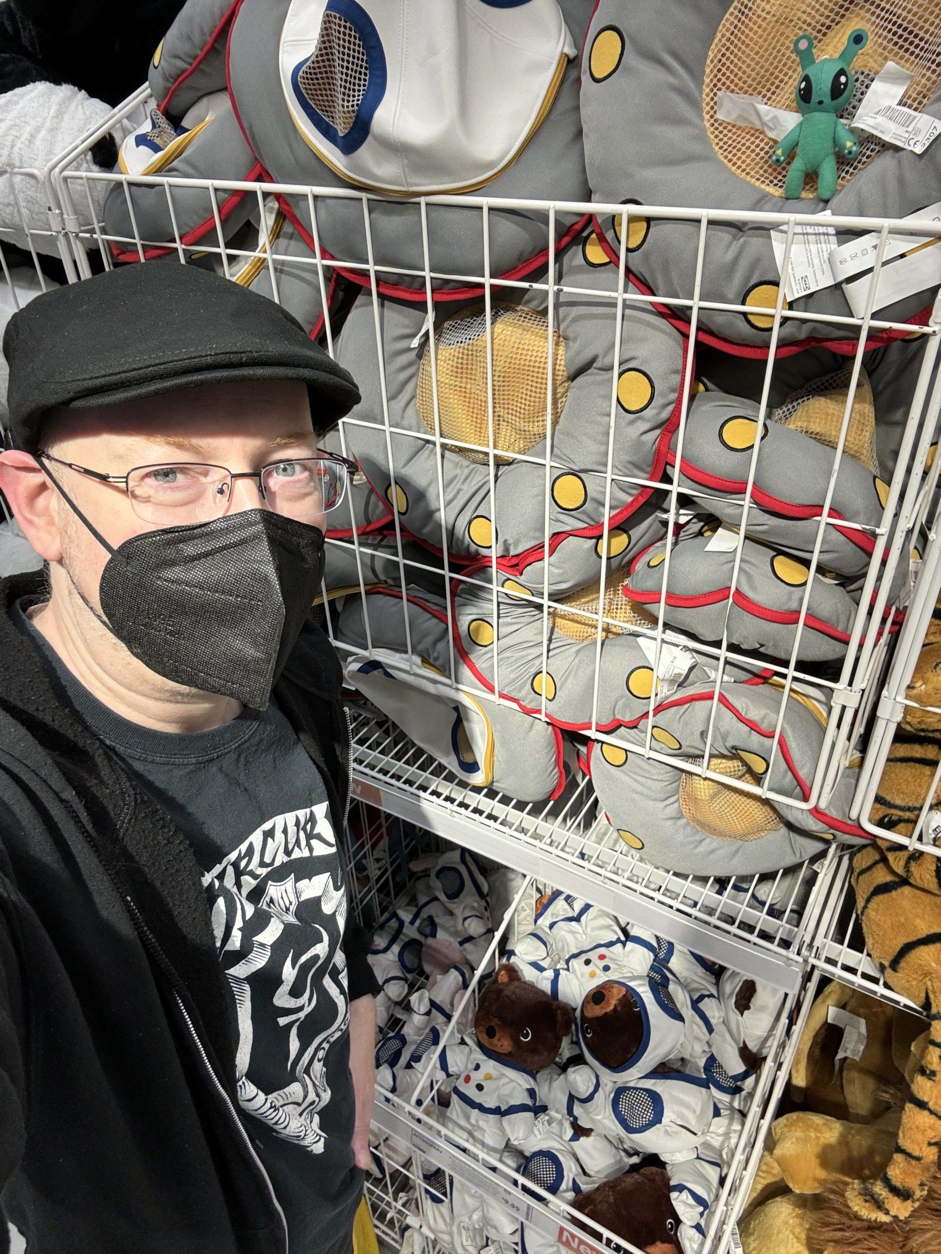 Me in front of IKEA wire racks of stuffed toys, including teddy bears in space suits and UFOs, and with a little green alien toy on one of the UFOs.