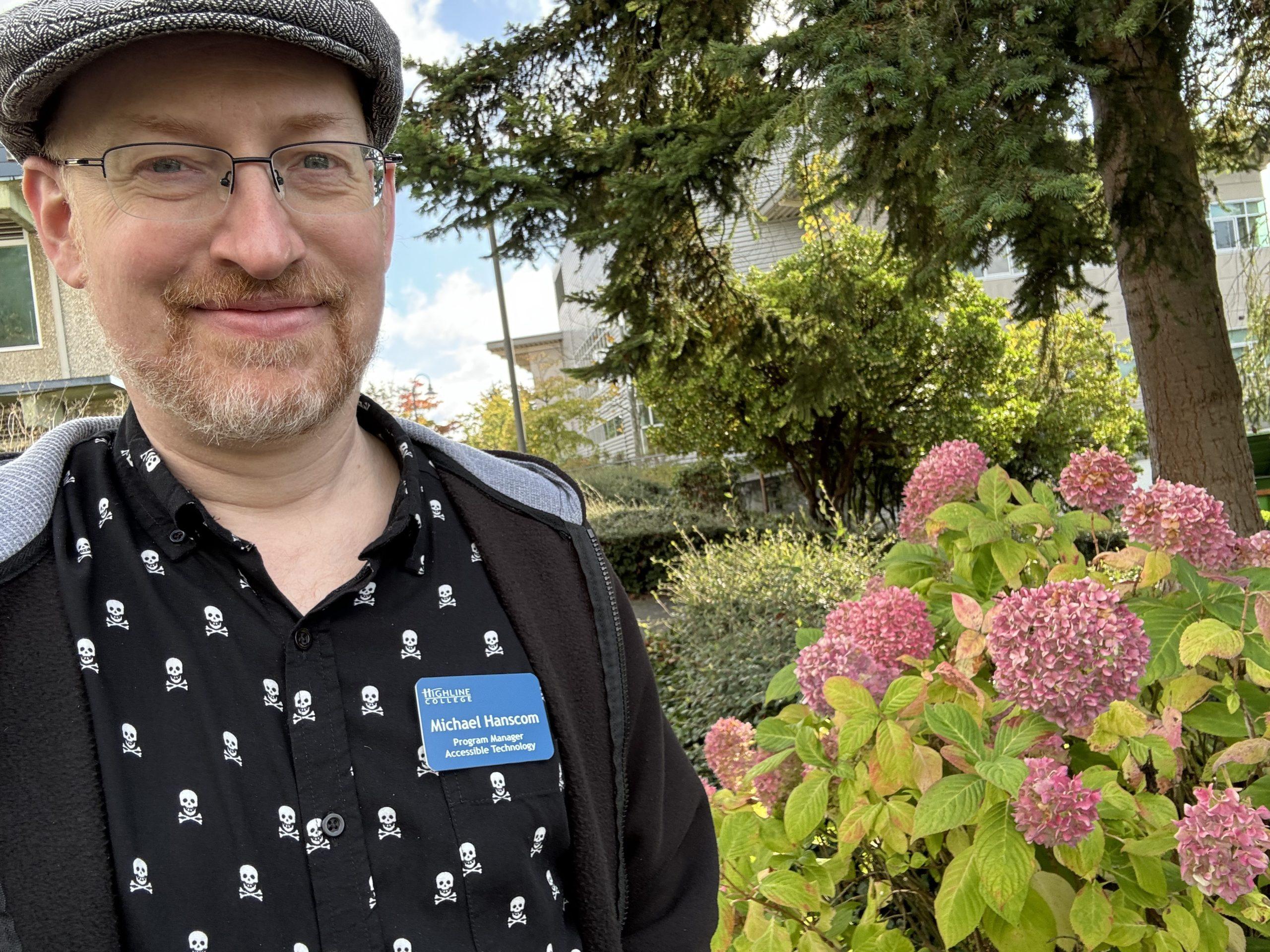 Me standing outside in front of some pink-flowered bushes, wearing a black hoodie over a black button-up shirt with a pattern of white skulls and crossbones.