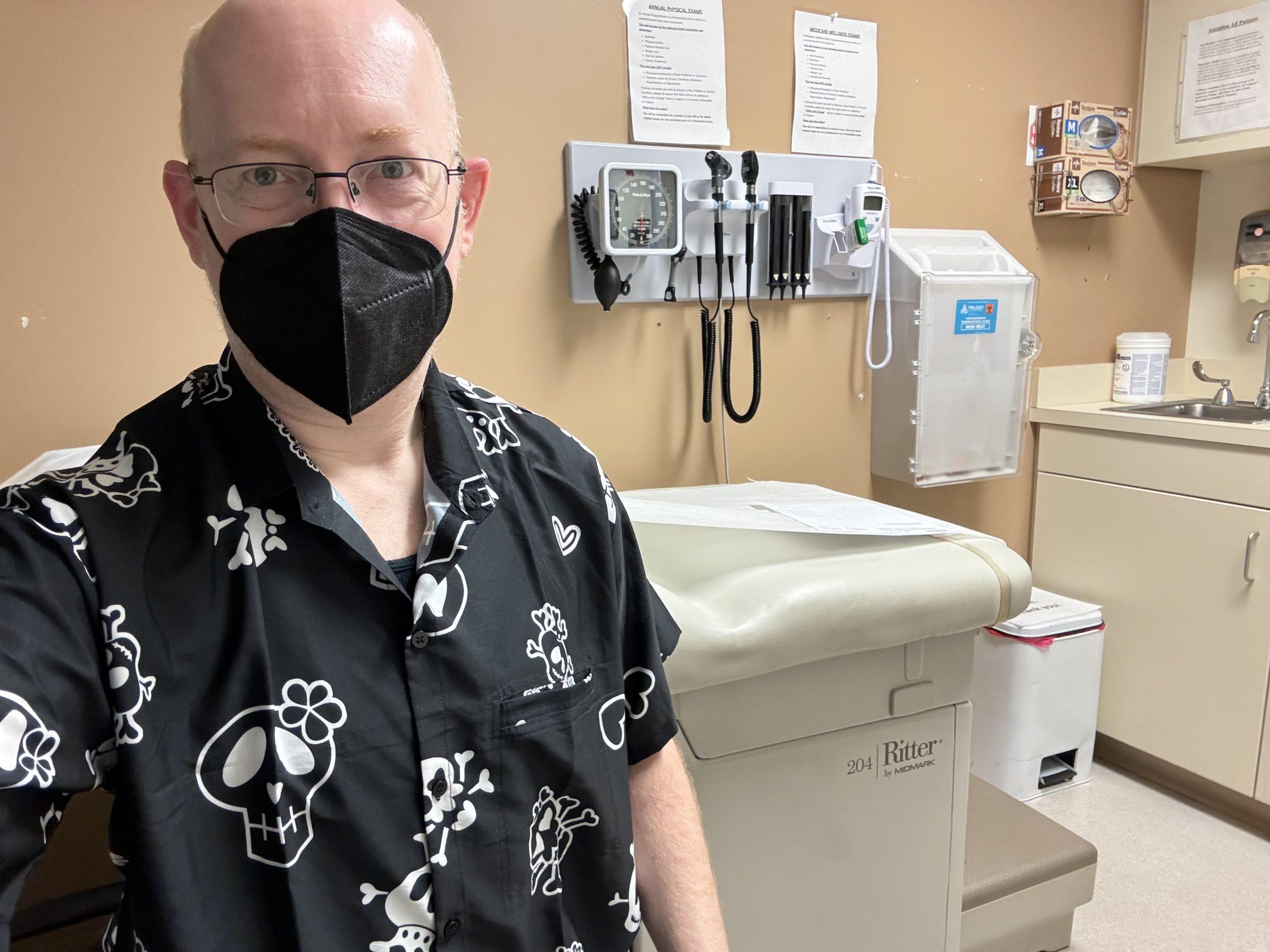 Me in a hospital examination room, masked, and wearing a black button-up short-sleeve shirt with large cute cartoon skulls on it.