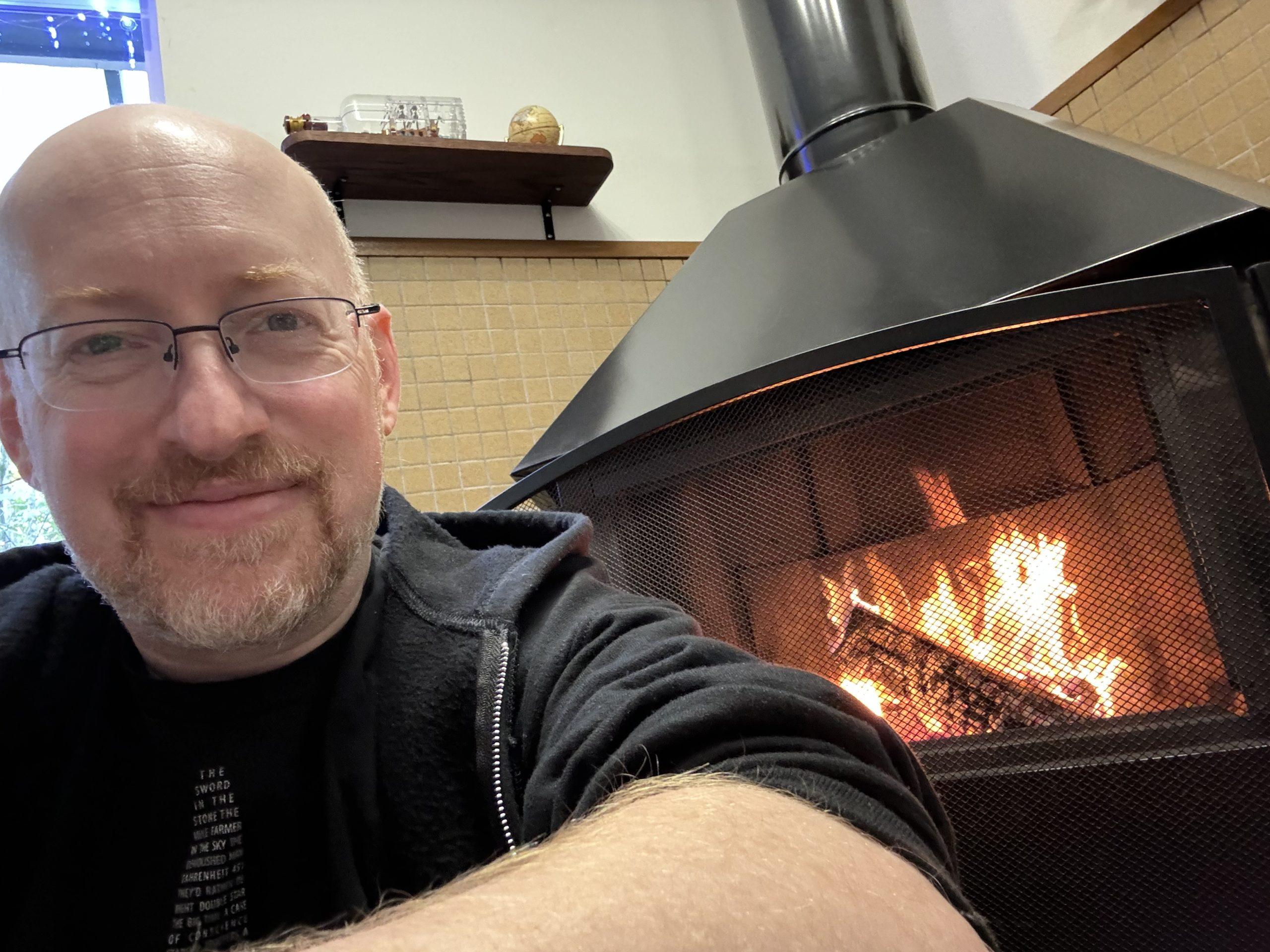 Me sitting in front of a black metal free-standing fireplace in the corner of our living room; logs are burning in the fireplace.