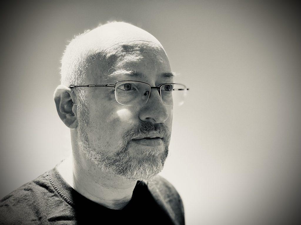 A black-and-white selfie of me, a white man with very short cropped hair and greying beard, wearing silver half-rim eyeglasses and a black t-shirt, looking off to the right of the photo with a neutral expression.