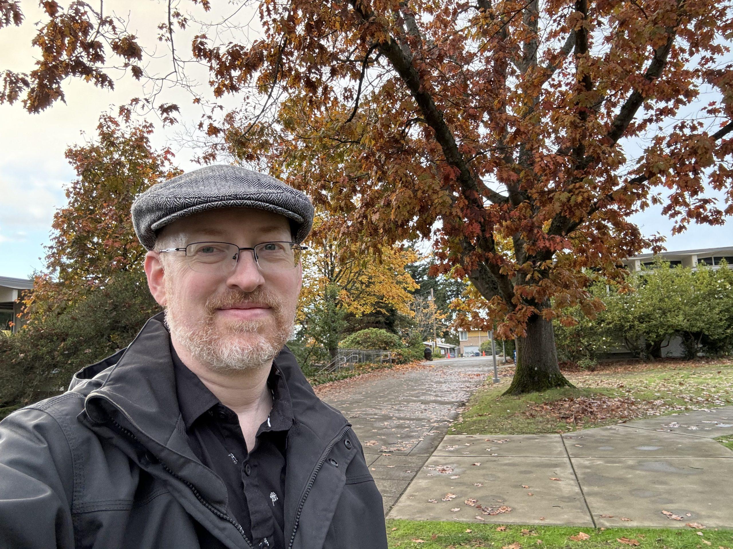 Me standing outside in front of a large tree with orangey-red fall leaves, with other trees in fall colors in the background, and lots of leaves on the ground and scattered across the wet pavement of a walkway.