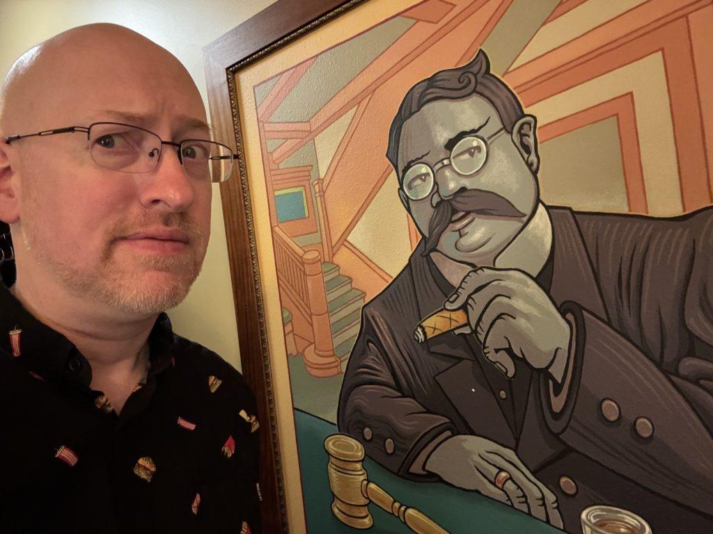 Me with a skeptical expression, next to a painting of a man who looks very much like, but is not, Teddy Roosevelt, smoking a cigar at a bar, with a drink and a gavel in front of him.