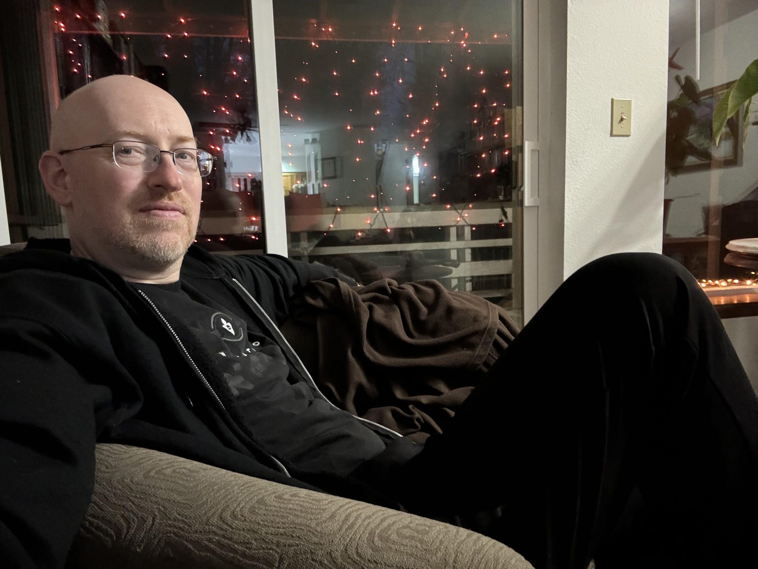 Me sitting in a living room chair, with orange twinkle lights visible through the sliding glass door behind me.