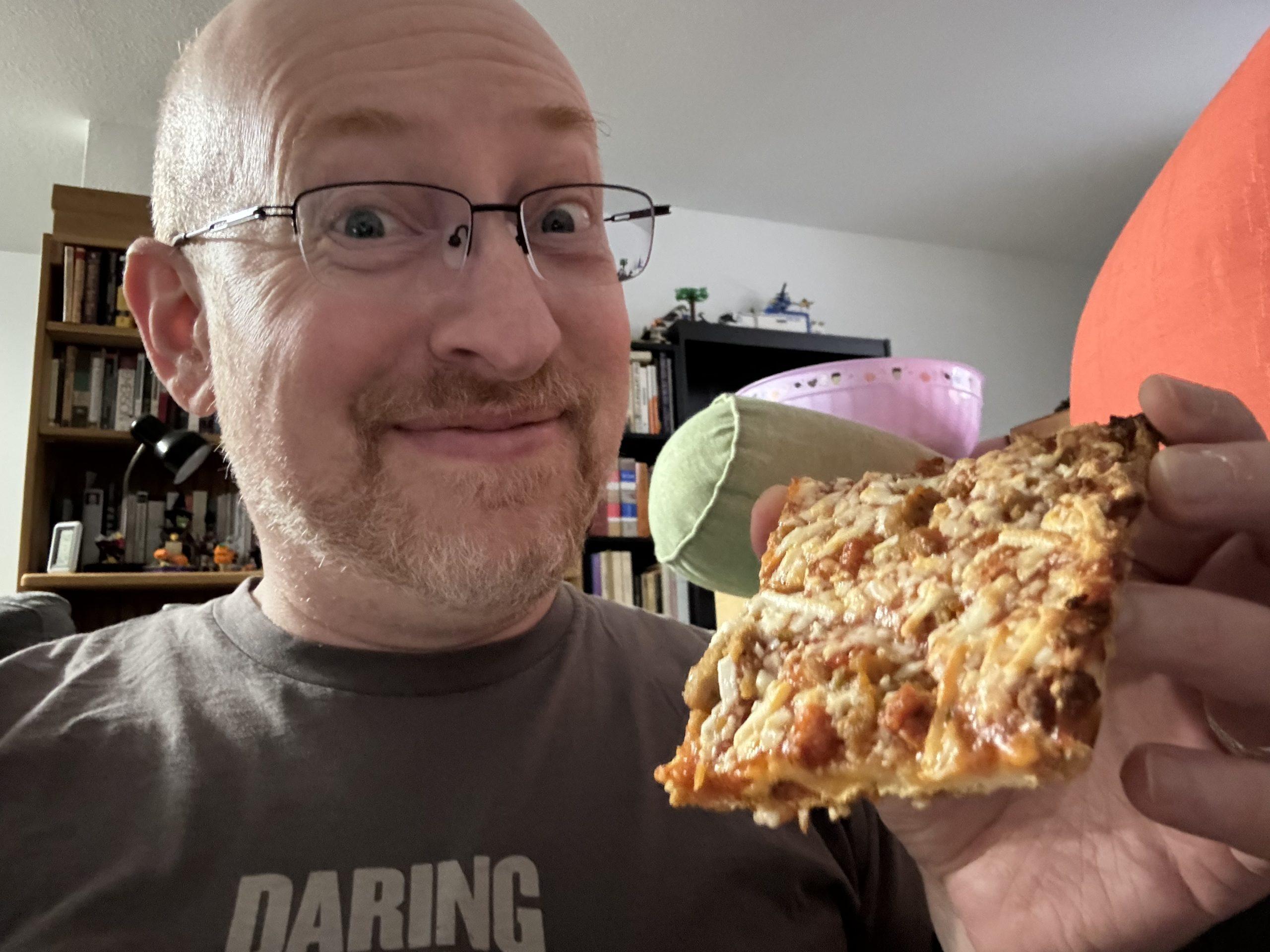 Me with a goofy smile and raised eyebrows, holding a piece of Tortino's pizza.