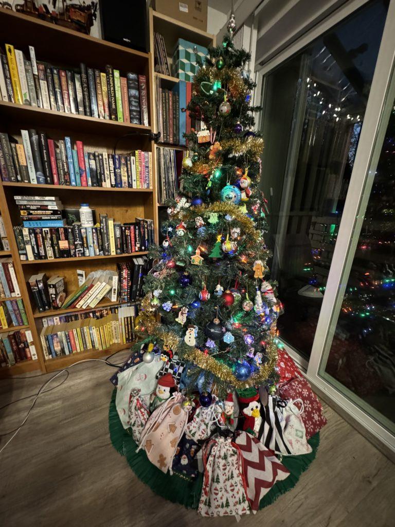 A Christmas tree in a living room corner, in front of a full bookcase on one side and a glass sliding door on the other. Gifts are piled underneath the tree.