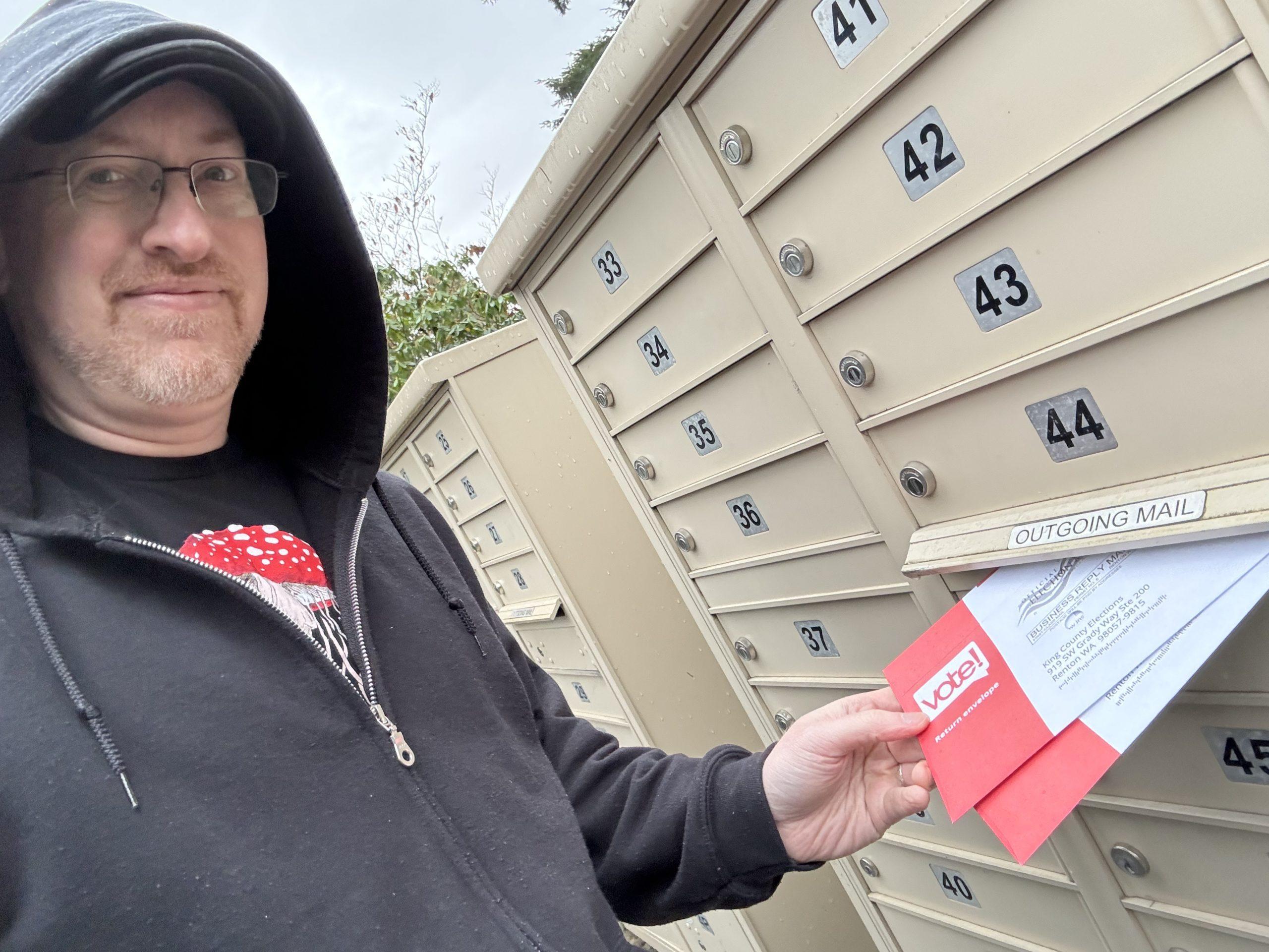 Me standing outside by a bank of mailboxes, putting two ballot envelopes into the outgoing mail slot.