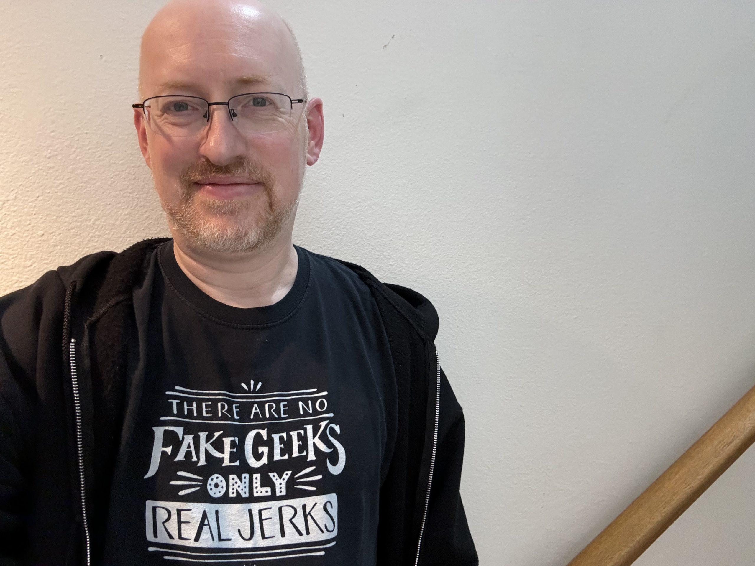 Me against a plain wall, wearing a t-shirt that says 'There are no fake geeks, only real jerks'.