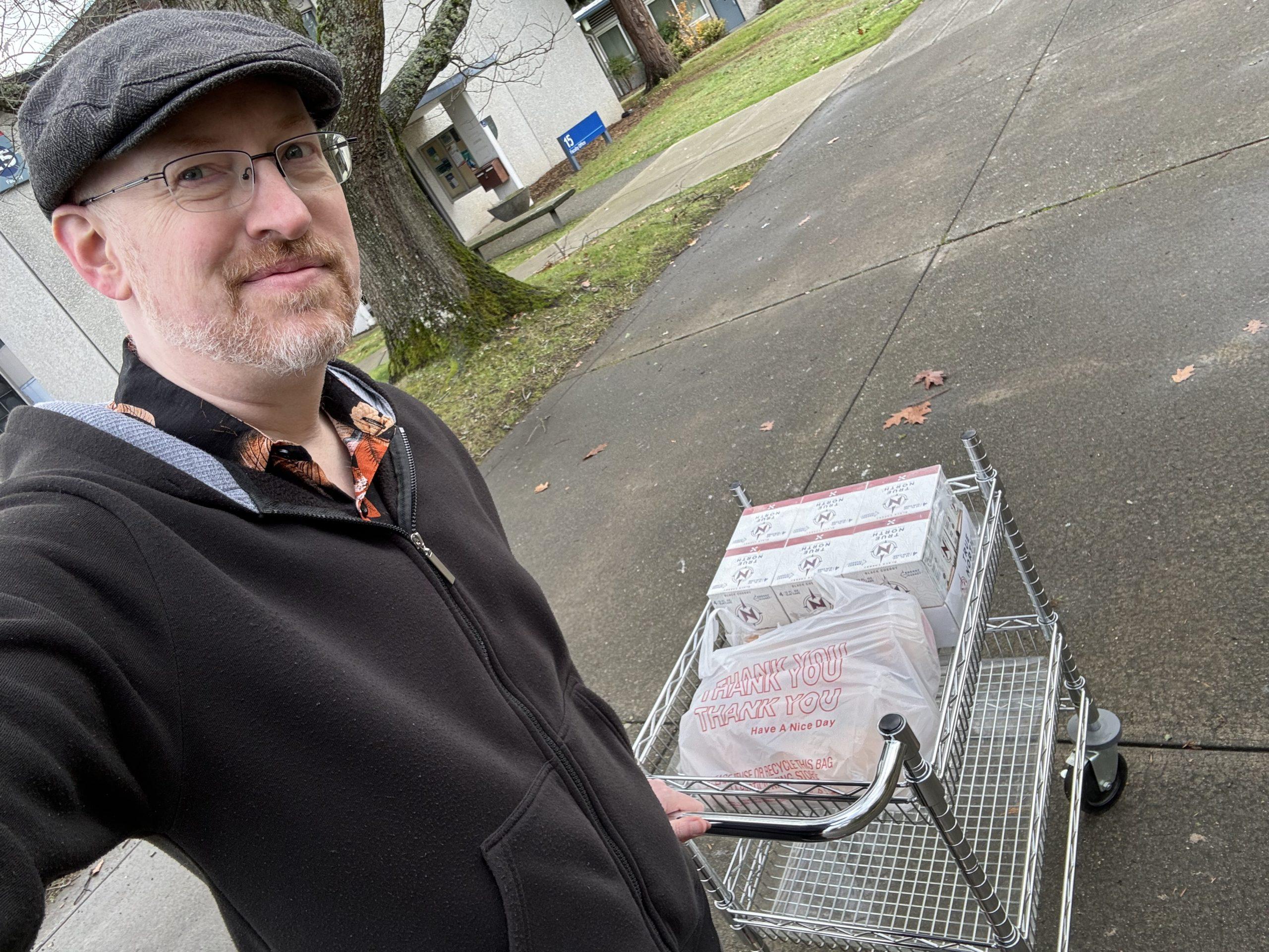Me outside on a sidewalk, pushing a metal cart with a grocery bag of food and a small case of sodas.