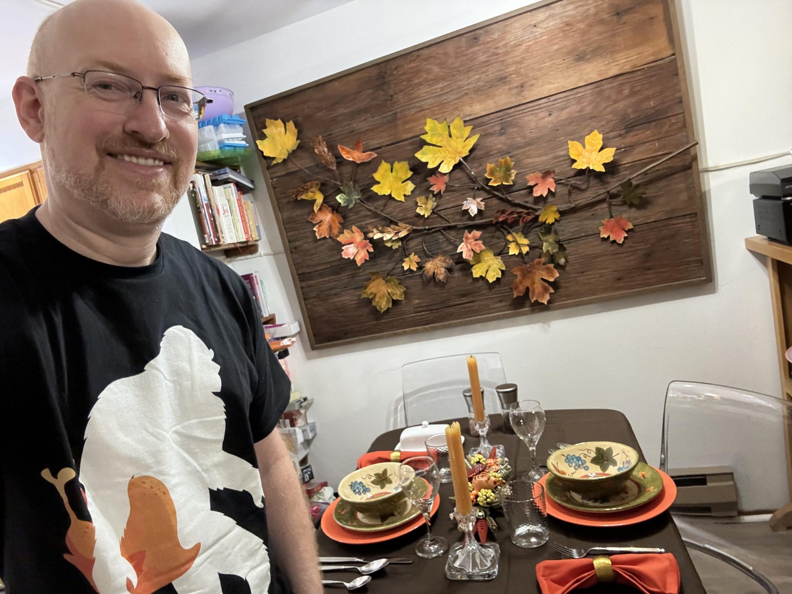Me in front of a table set for dinner with fall-colored table settings (brown tablecloth, orange dinner plates and napkins, green salad plates and bowls) and yellow candles. There is sculptural artwork on the wall of fall leaves mounted on a wood backing. I'm wearing a black t-shirt with a simple design of Bigfoot carrying a turkey.
