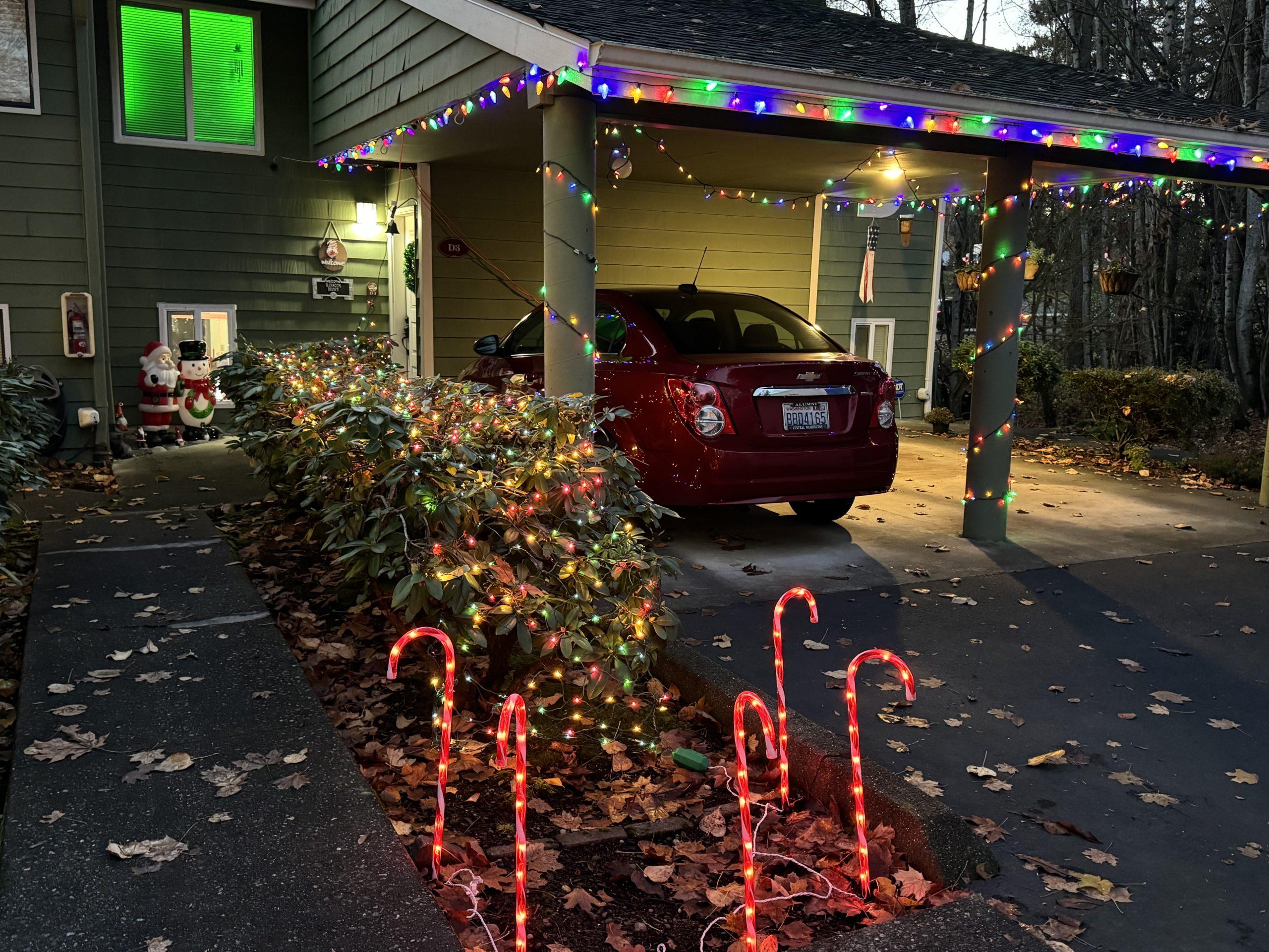 The front of our house, 1/4 of a condo fourplex, with colored holiday lights strung around the carport and over bushes by a walkway, lit candy canes in front of the bush, and Santa and snowman figures near the front door.