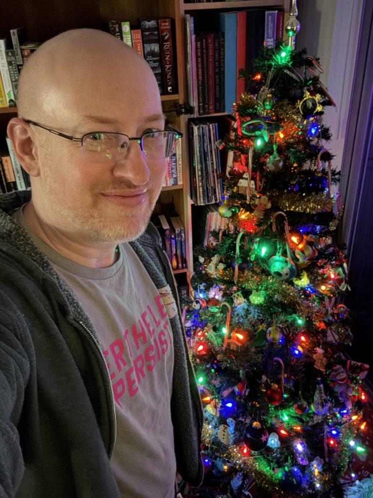 Me in a dim room, standing in front of a Christmas tree decorated with ornaments, candy canes, and multicolored lights.