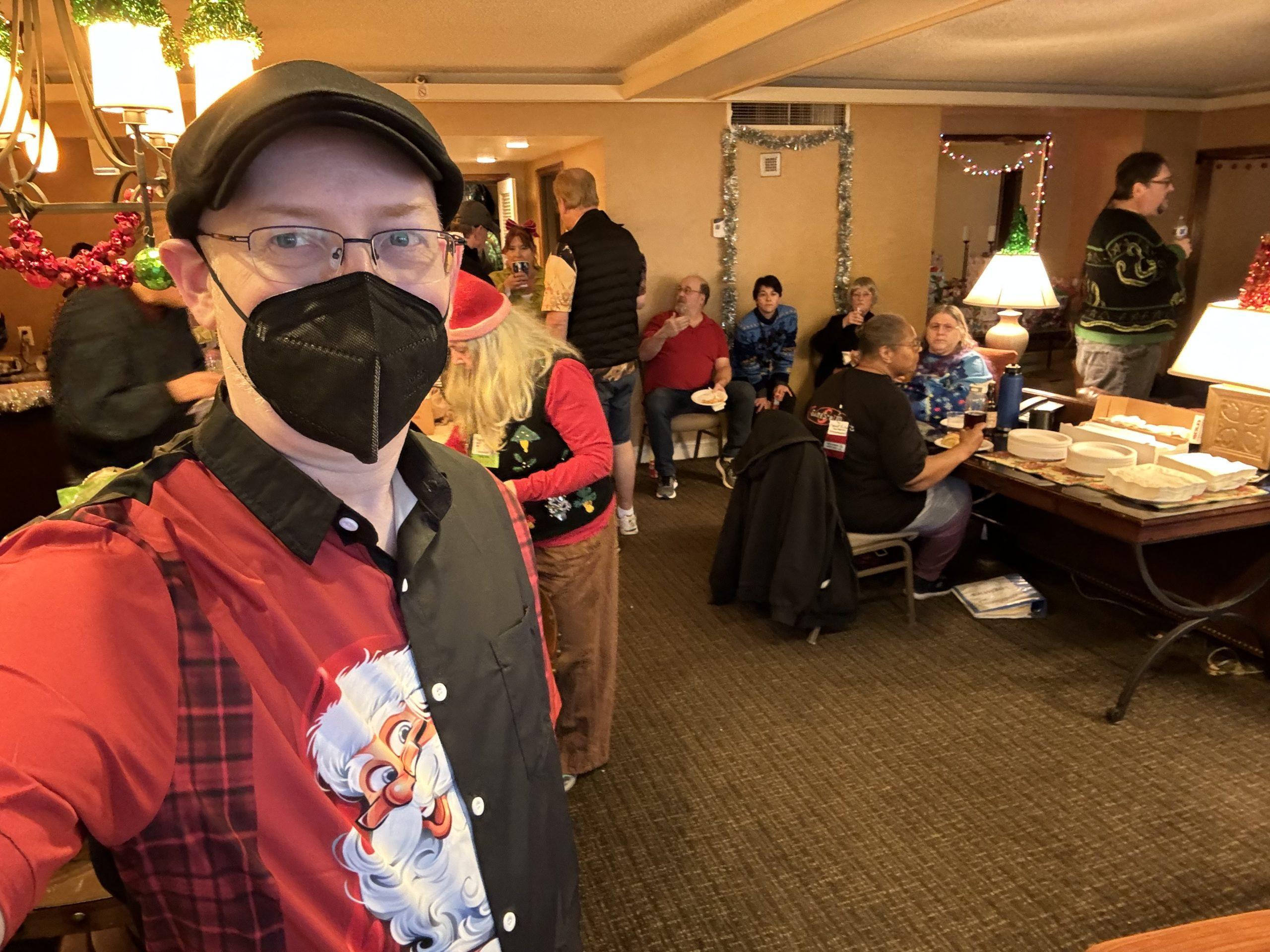 Me, wearing a mask and a button-up shirt that’s black on one side and has Santa peeking out from behind the buttons on the other, in a hotel room with holiday decorations and people behind me eating and chatting.