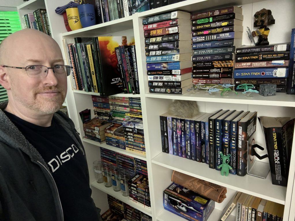 Me wearing a “Disco” Star Trek Discovery t-shirt, standing in front of shelves filled with Star Trek books.