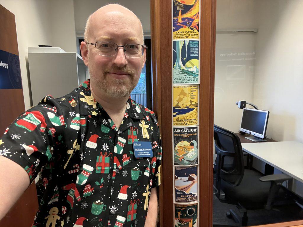 Me in my office at work, standing next to a door jamb decorated with a series of National Park style postcards advertising various places in the solar system. I’m wearing a black short-sleeve button-up shirt with holiday gifts, Santa hats, gingerbread people, candy canes, mittens, hot chocolate mugs, and snowflakes.