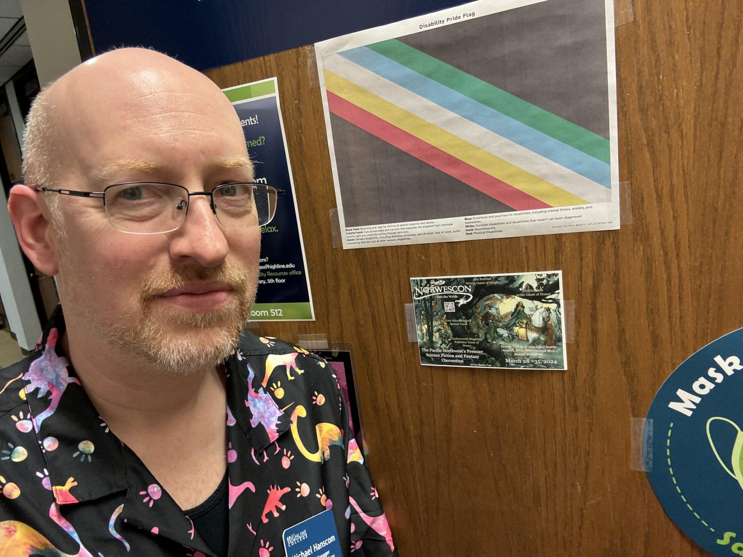 Me in front of my office door at work. A Norwescon postcard ad is taped to the door just underneath a disability pride flag.