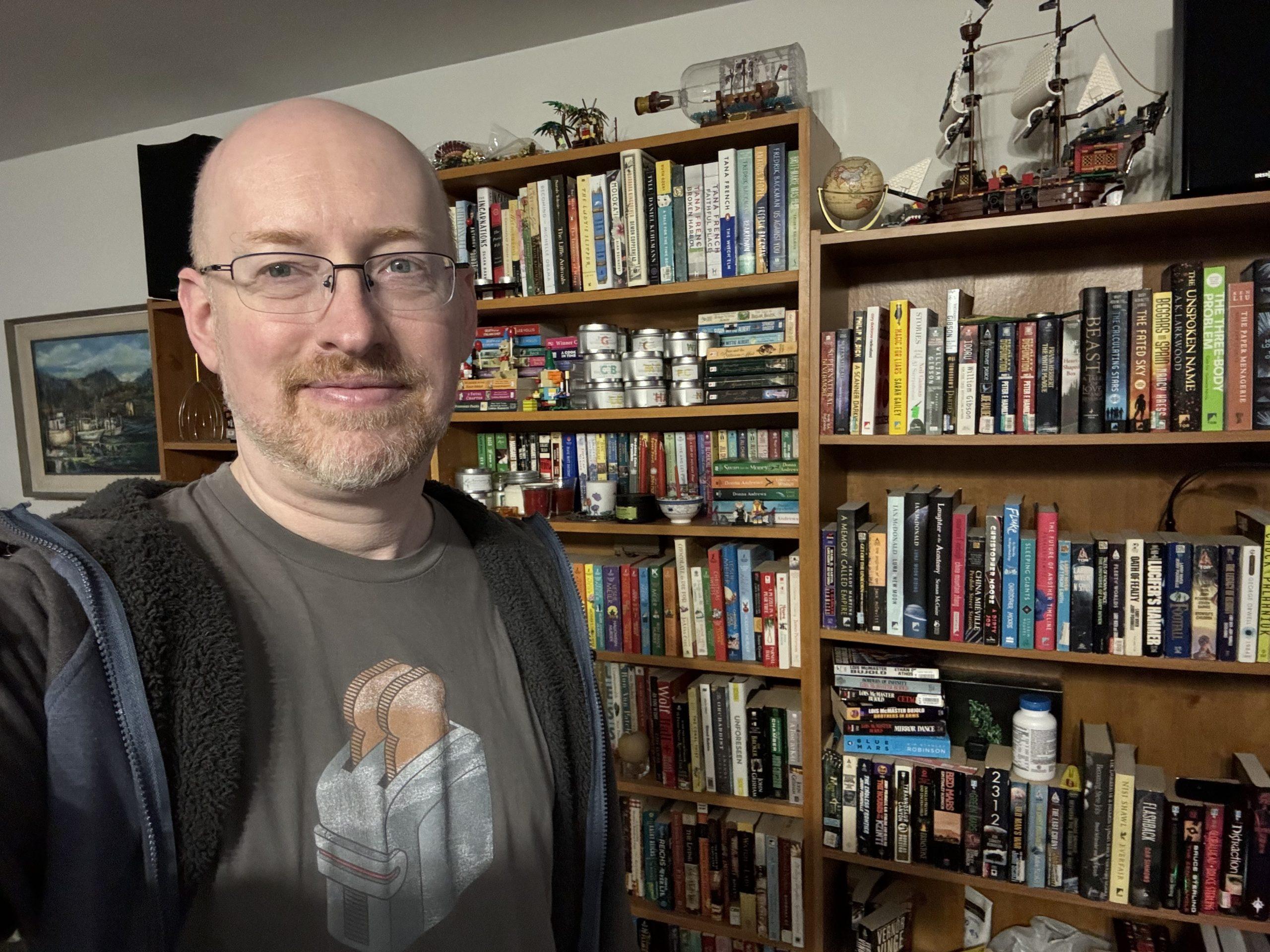 Me standing in front of a couple bookcases filled with books. A Lego sailing ship and ship-in-a-bottle are on display on top of the shelves.