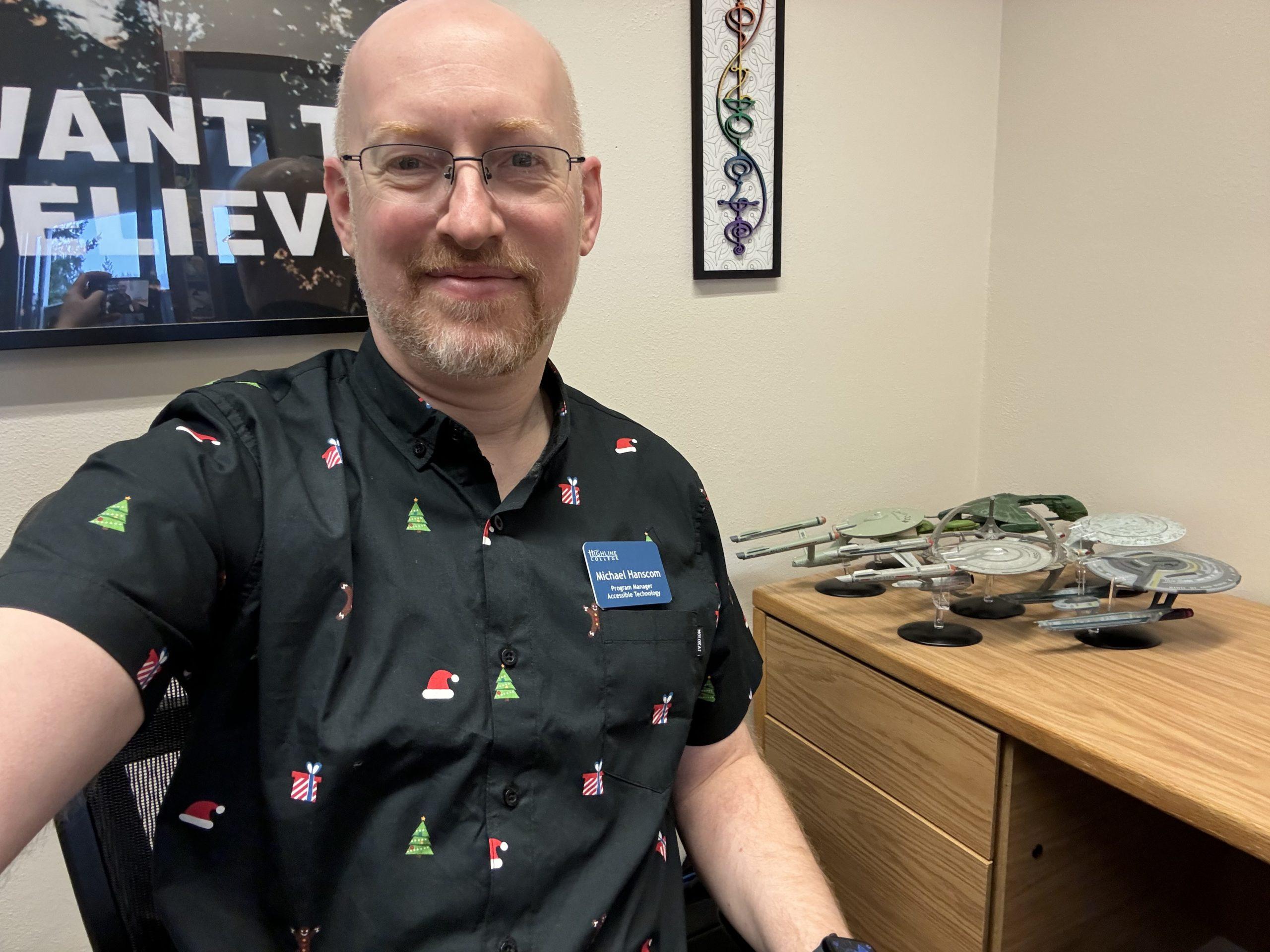 Me at my desk at work, wearing a black short-sleeve button-up shirt with santa hats, holiday trees, and presents on it.