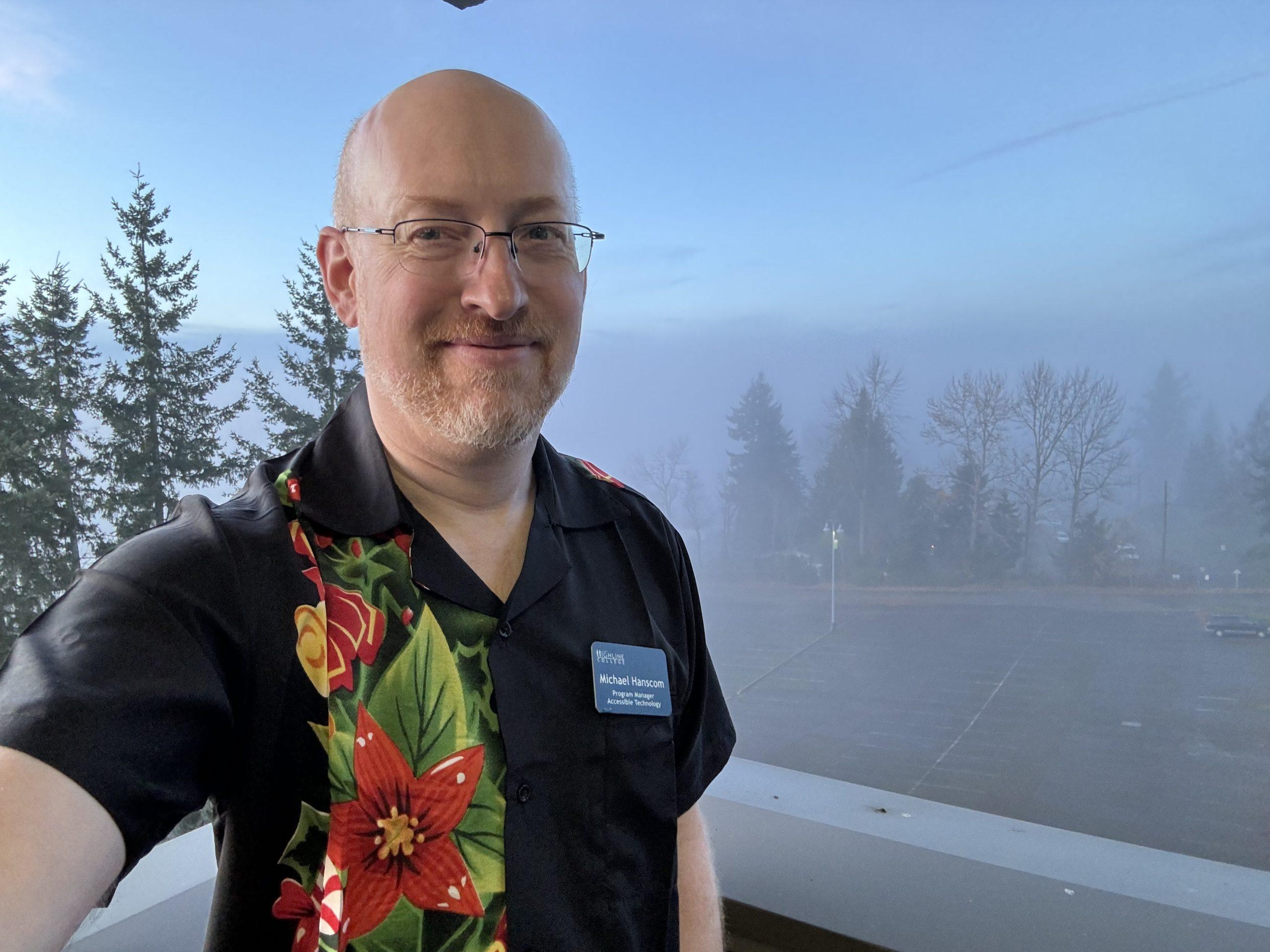 Me wearing a black short-sleeve button-up shirt with colorful poinsettas, holly, and candy canes on the right side of the shirt; behind me is a slightly-visible parking lot shrouded in fog.