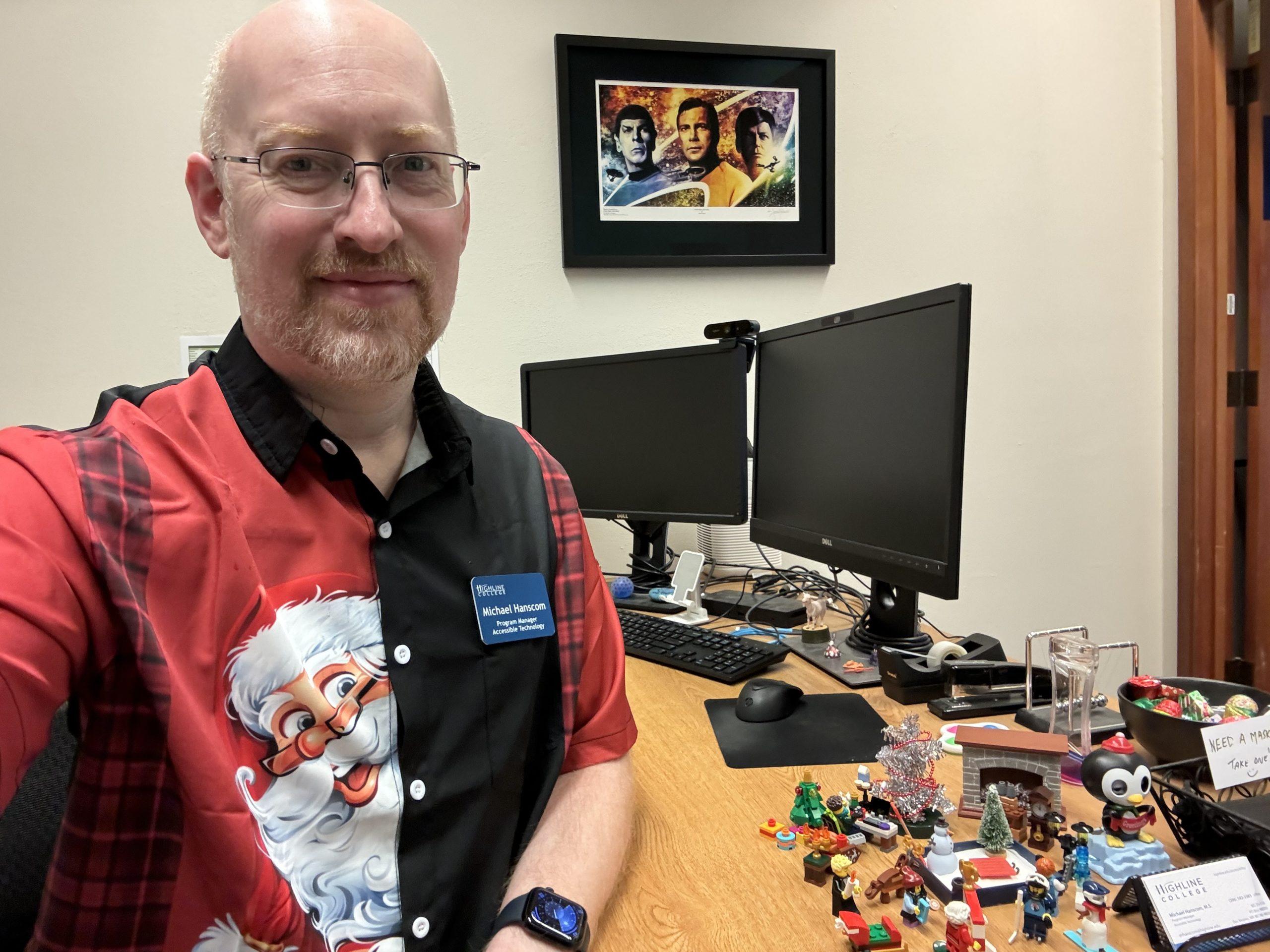 Me in my work office, wearing a short-sleeve button-up shirt that's black on my left, red on my right, with plaid sides and red sleeves, and with Santa peeking out from behind the buttons. Visible on my desk are a group of holiday themed toys.