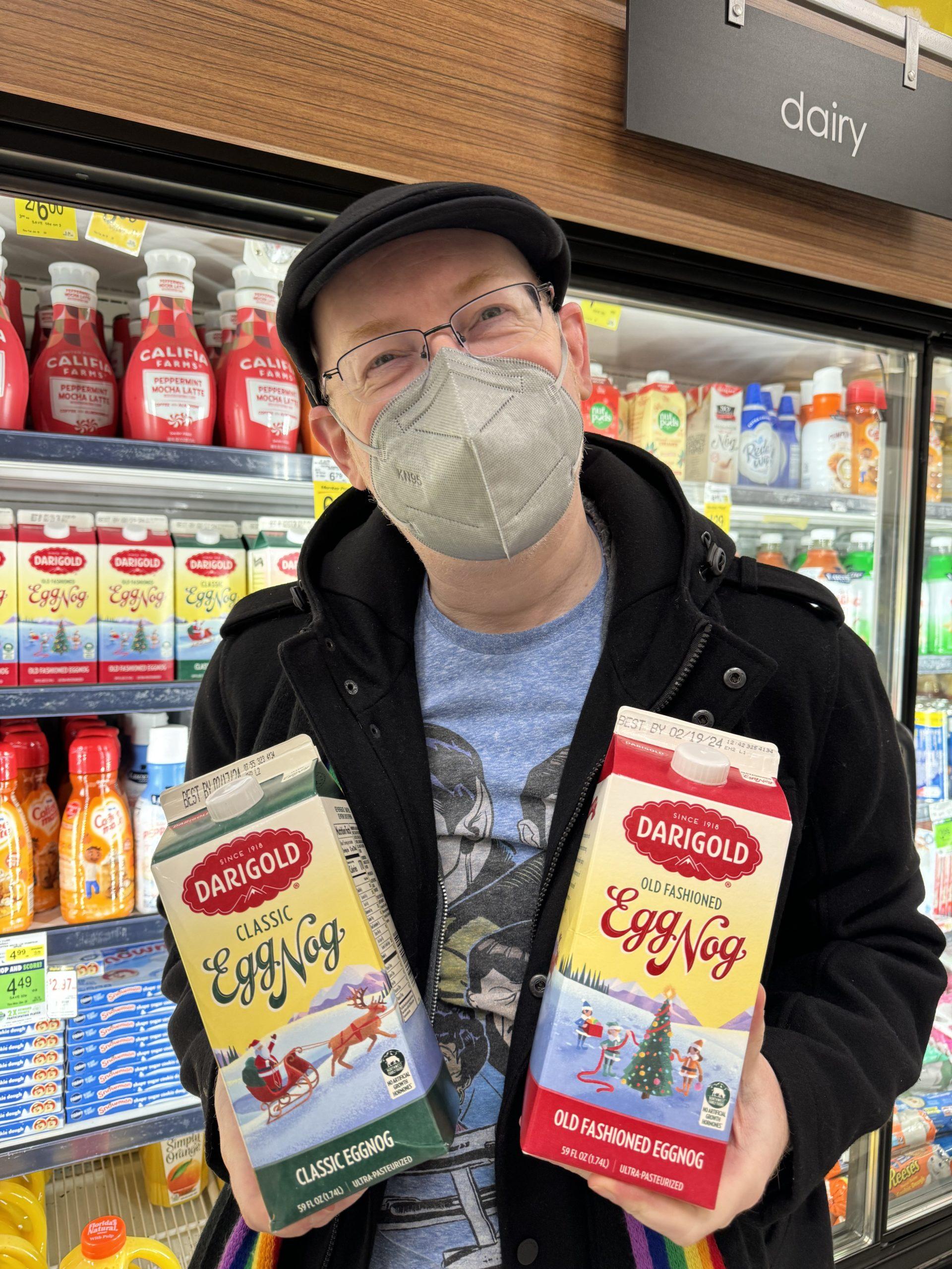 Me (masked) in a grocery store, with a confused expression and holding two cartons of Darigold egg nog, one labeled 'classic' and one labeled 'old fashioned'.