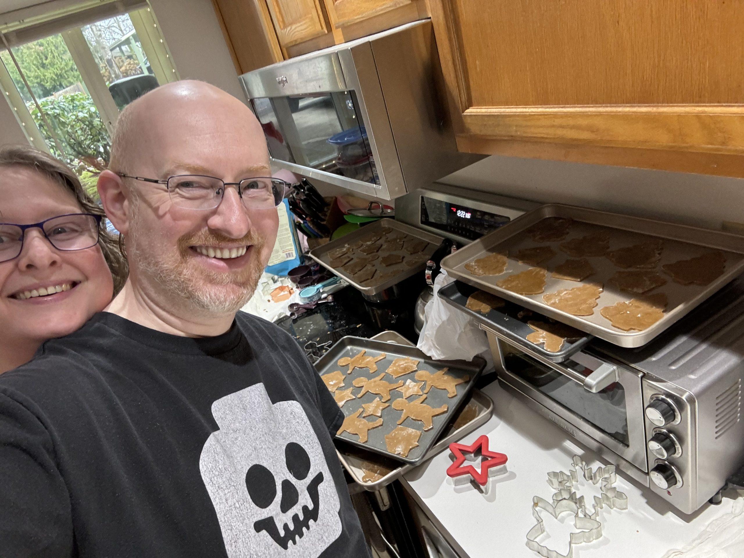 Me in our kitchen, with several baking sheets of gingerbread cookies ready to go in the oven on the counter. My wife is peeking over my shoulder and smiling.