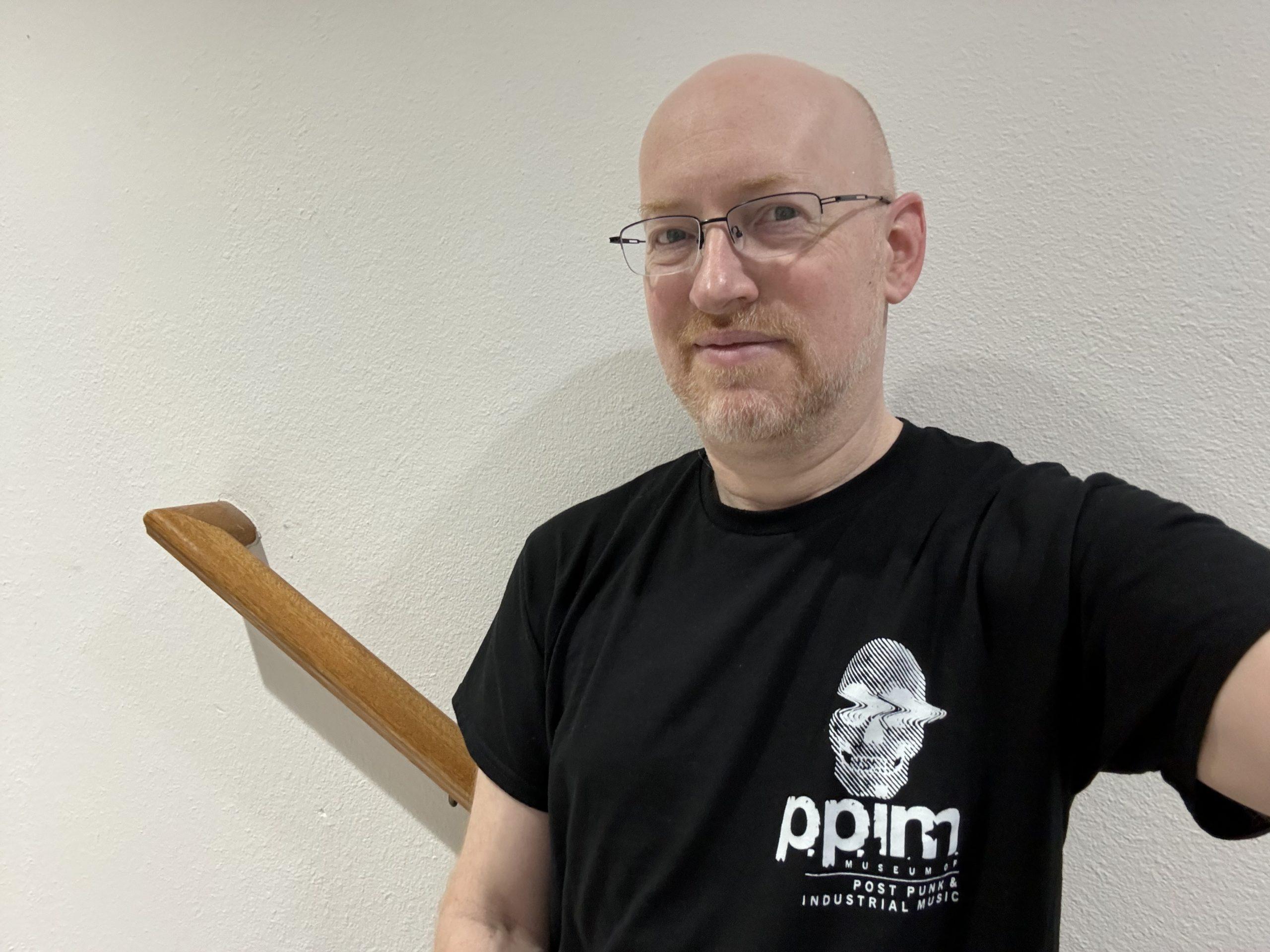 Me standing against a blank wall, wearing a black t-shirt with the logo for the Museum of Post-Punk and Industrial Music.