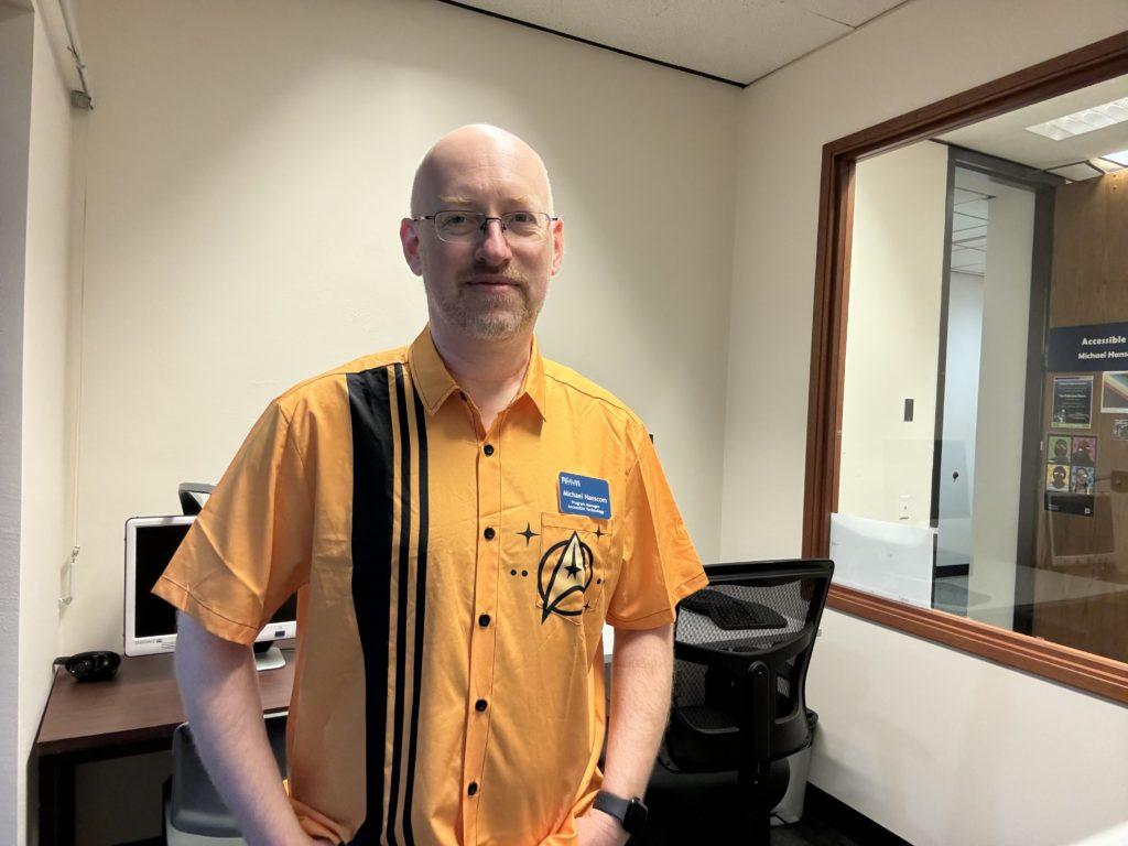 Me in my work office, wearing a retro bowling shirt style button up in TOS Starfleet gold, with the delta shield logo on the breast pocket, and three black stripes running down the right side.