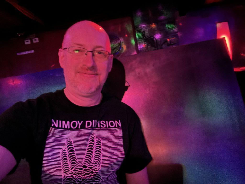 Me, in red/pnk/purple lighting, sitting on a booth in a goth club, wearing a t-shirt with a parody of the Joy Division ‘Unknown Pleasures’ art that says ‘Nimoy Division’ with the squiggles forming the Vulcan salute.