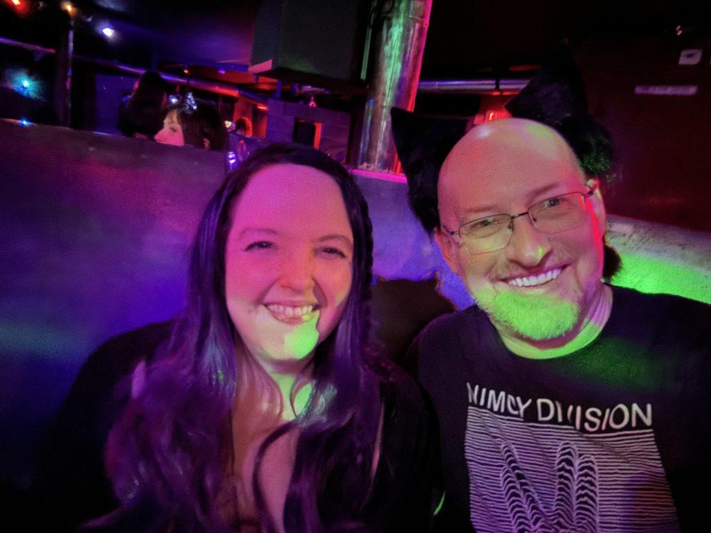 Me, in red/pnk/purple lighting, sitting on a booth in a goth club, next to Michelle, a white woman with long dark hair wearing a black top.