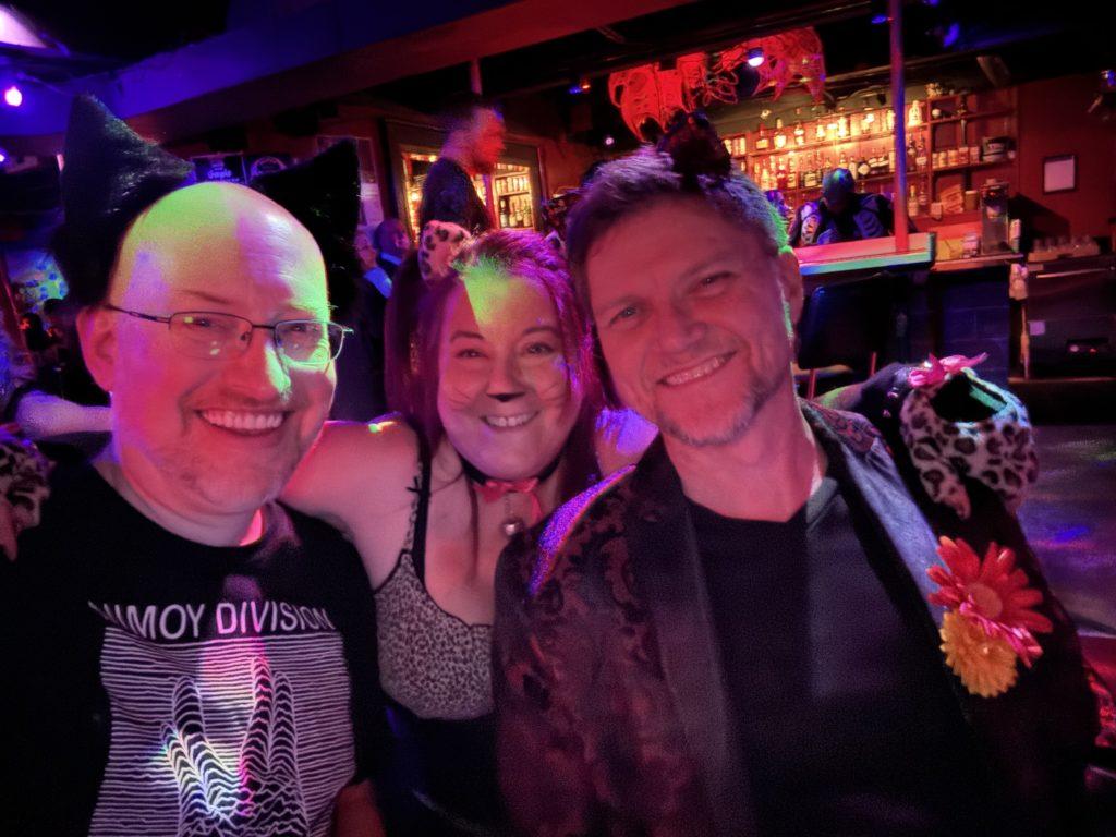 Me, in red/pnk/purple lighting, sitting on a booth in a goth club, next to Teya, a white woman with purple hair and cat-style face makeup, and her husband Jon, a white man with short brown hair wearing a dark floral jacket over a black shirt. Teya and I are both wearing cat ear headbands.
