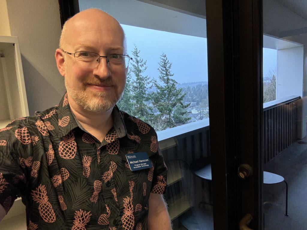 Me in my office at work, with a cold drizzly day visible through the window behind me, wearing a black shirt with pineapples printed on it.