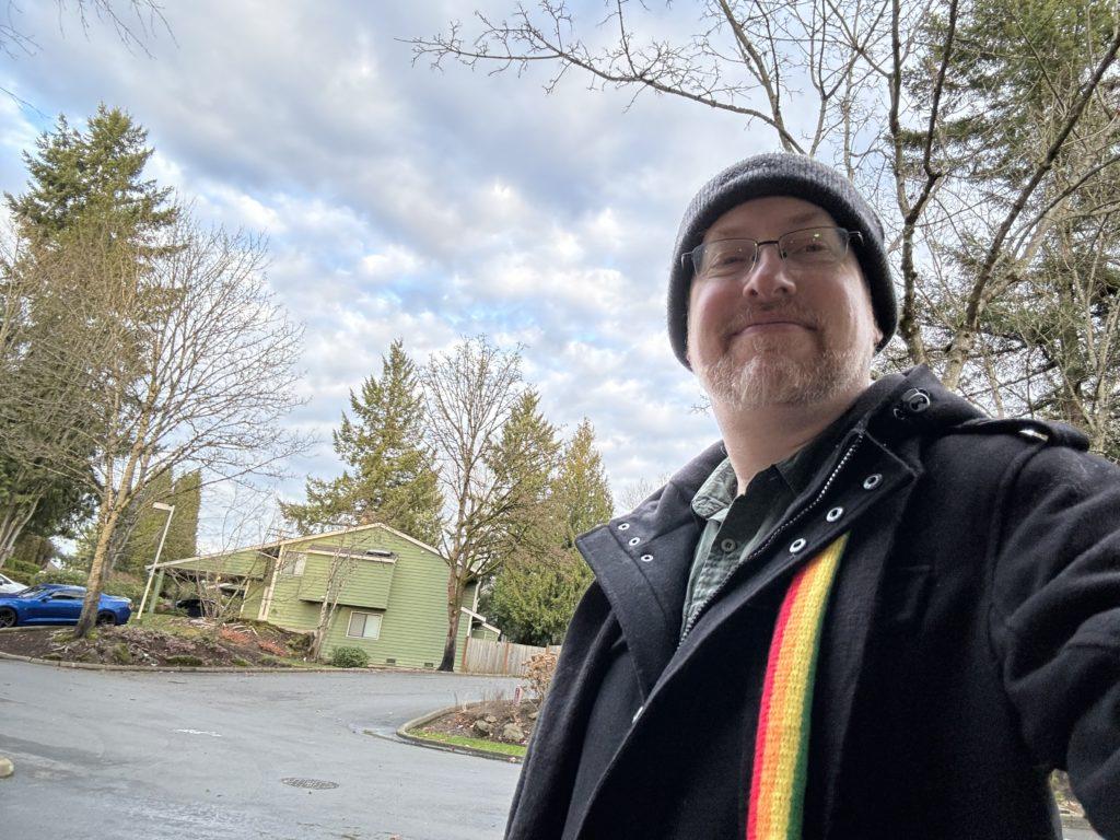 Me standing outside under a cloudy but slightly blue afternoon sky, wearing a black wool coat with rainbow scarf.