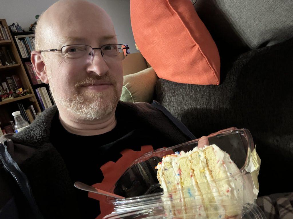 Me holding a clear plastic container with a large piece of grocery store cake (vanilla with vanilla frosting and colored sprinkles).