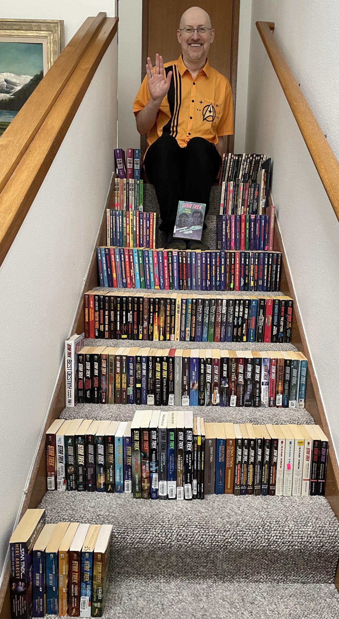 Me sitting at the top of a staircase, wearing a retro bowling shirt style shirt that&#39;s Star Trek gold and has the delta shield on the breast pocket. I&#39;m giving the Vulcan salute. Arrayed across the stairs to either side and below me are a lot of Star Trek books.