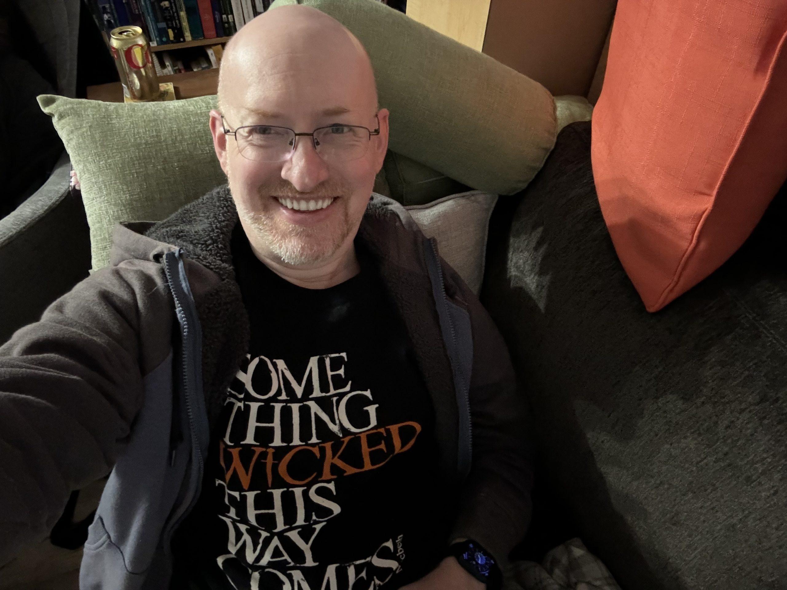 Me on our living room couch, wearing a t-shirt that says 'something wicked this way comes'.