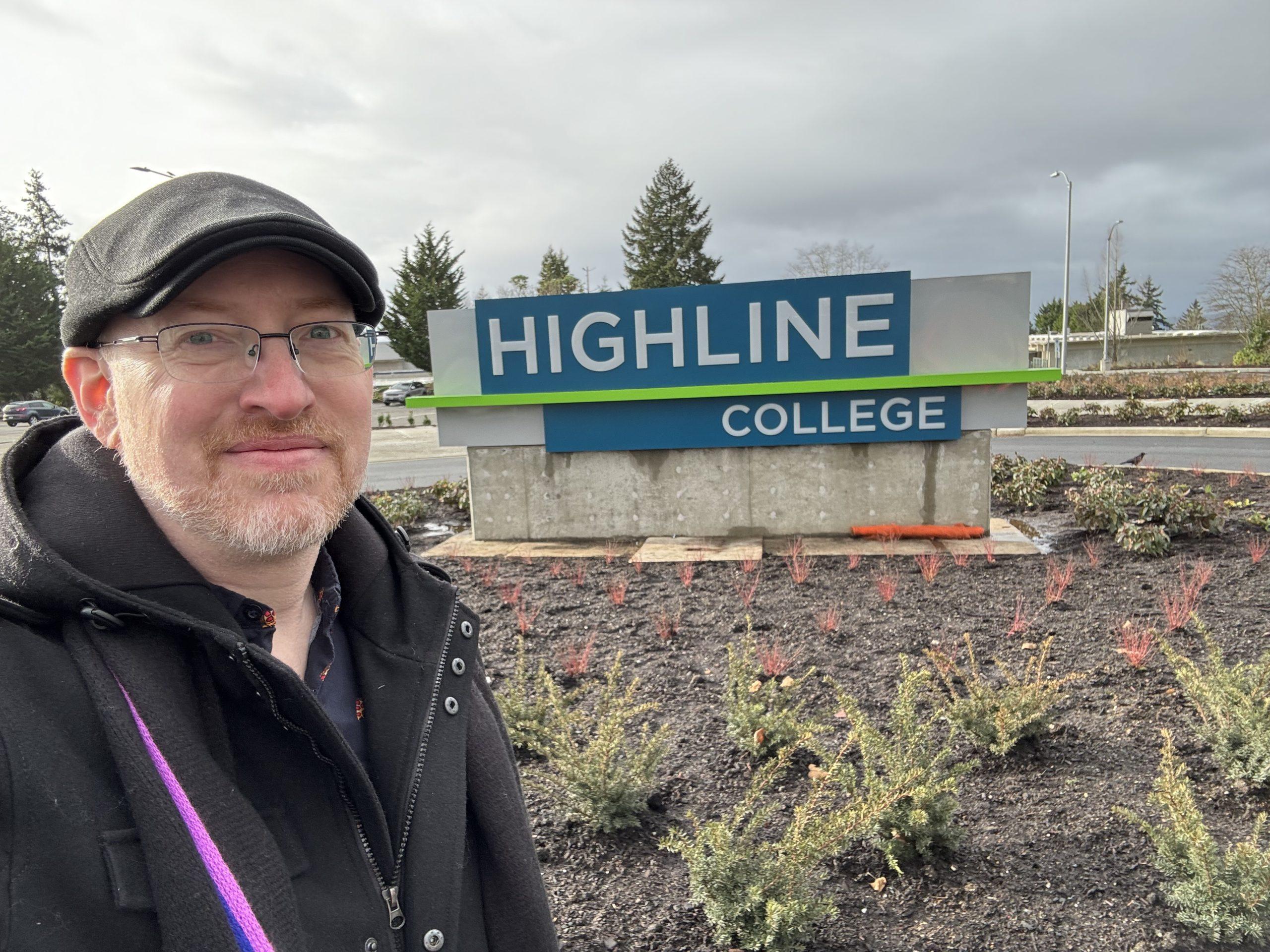 Me standing outside in front of the new front entrance sign for Highline College, a large sign in the center of a roundabout with just-planted plants around it.