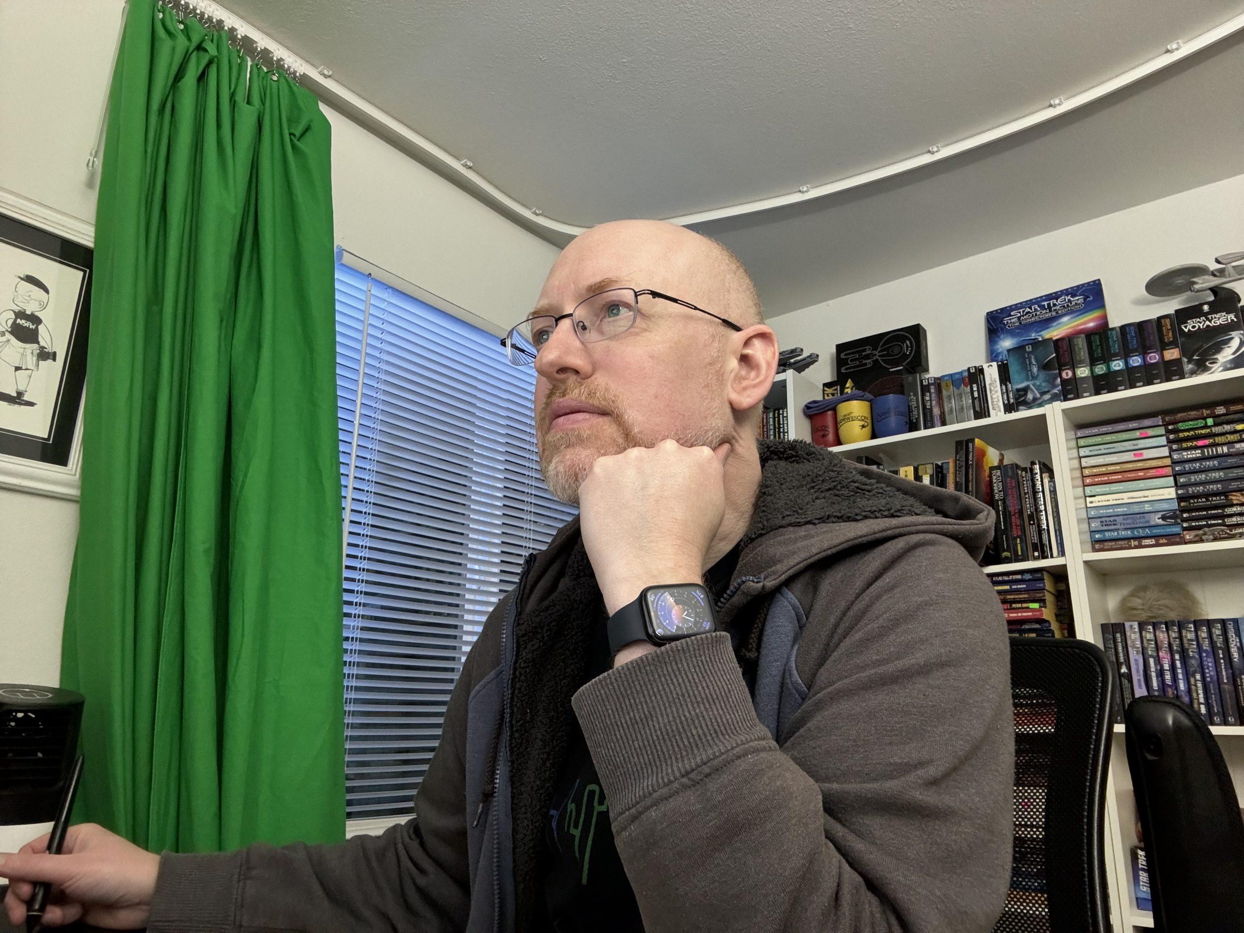 Me sitting in my home office, with a green screen curtain to my right and shelves of books behind me, looking at my computer.