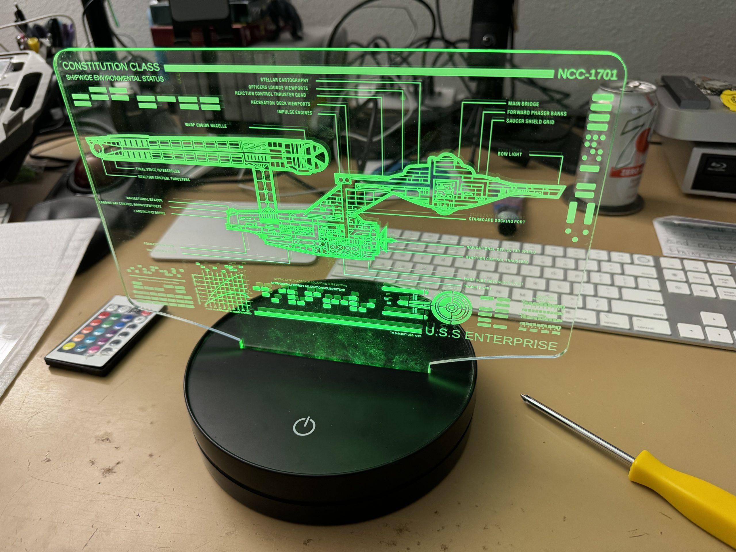 A circular black stands holds a clear panel with a schematic of the original USS Enterprise which is glowing green. Also visible near or through the panel is a small remote to change LED colors, a screwdriver, and the keyboard and trackpad for my computer.