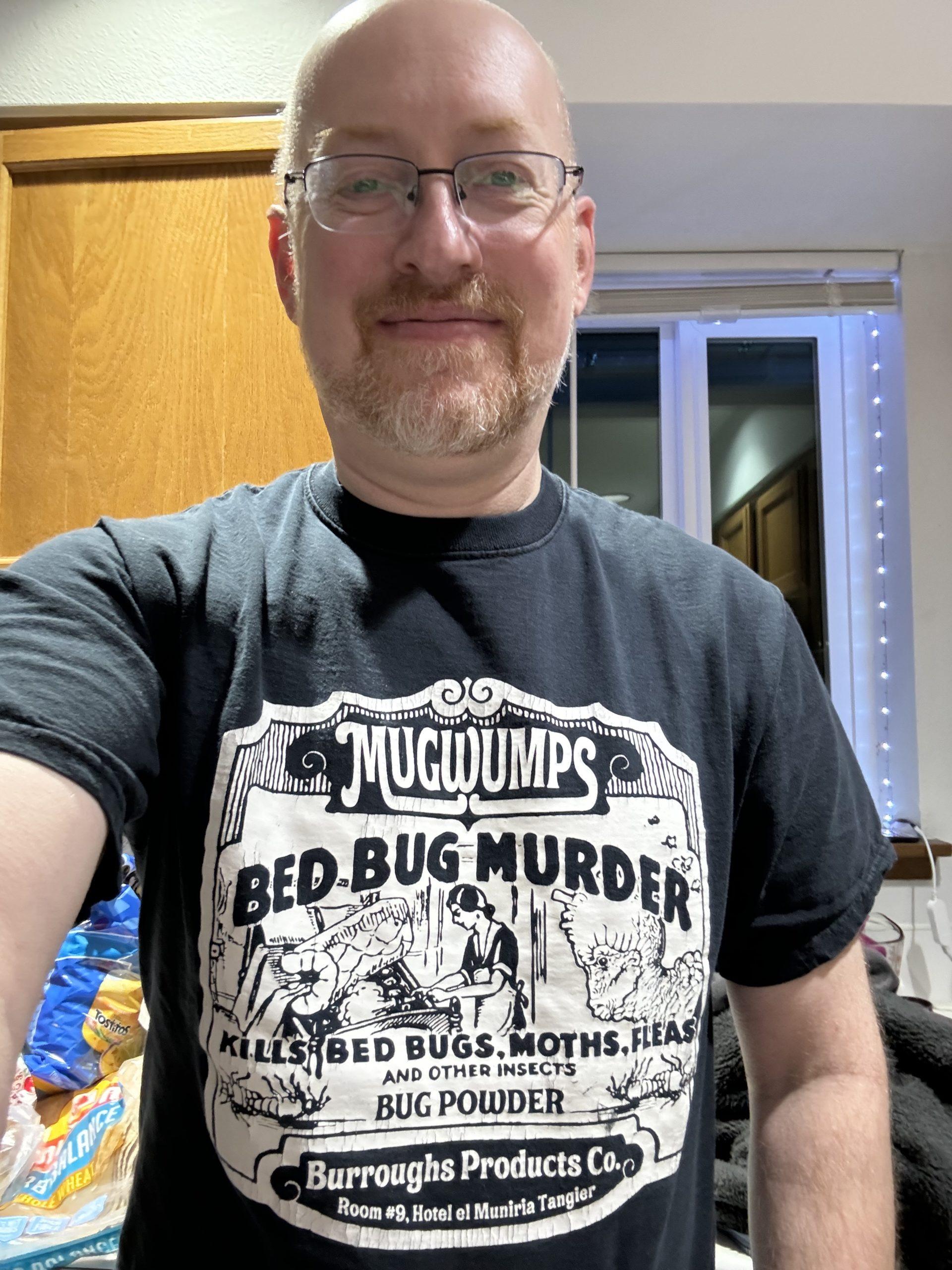 Me wearing a black t-shirt with a design that looks like an old-time label that says: Mugwumps Bed Bug Murder Bug Powder. Kills bed bugs, moths, fleas, and other insects. Burroughs Products Co. Room #9, Hotel el Muniria Tangier.