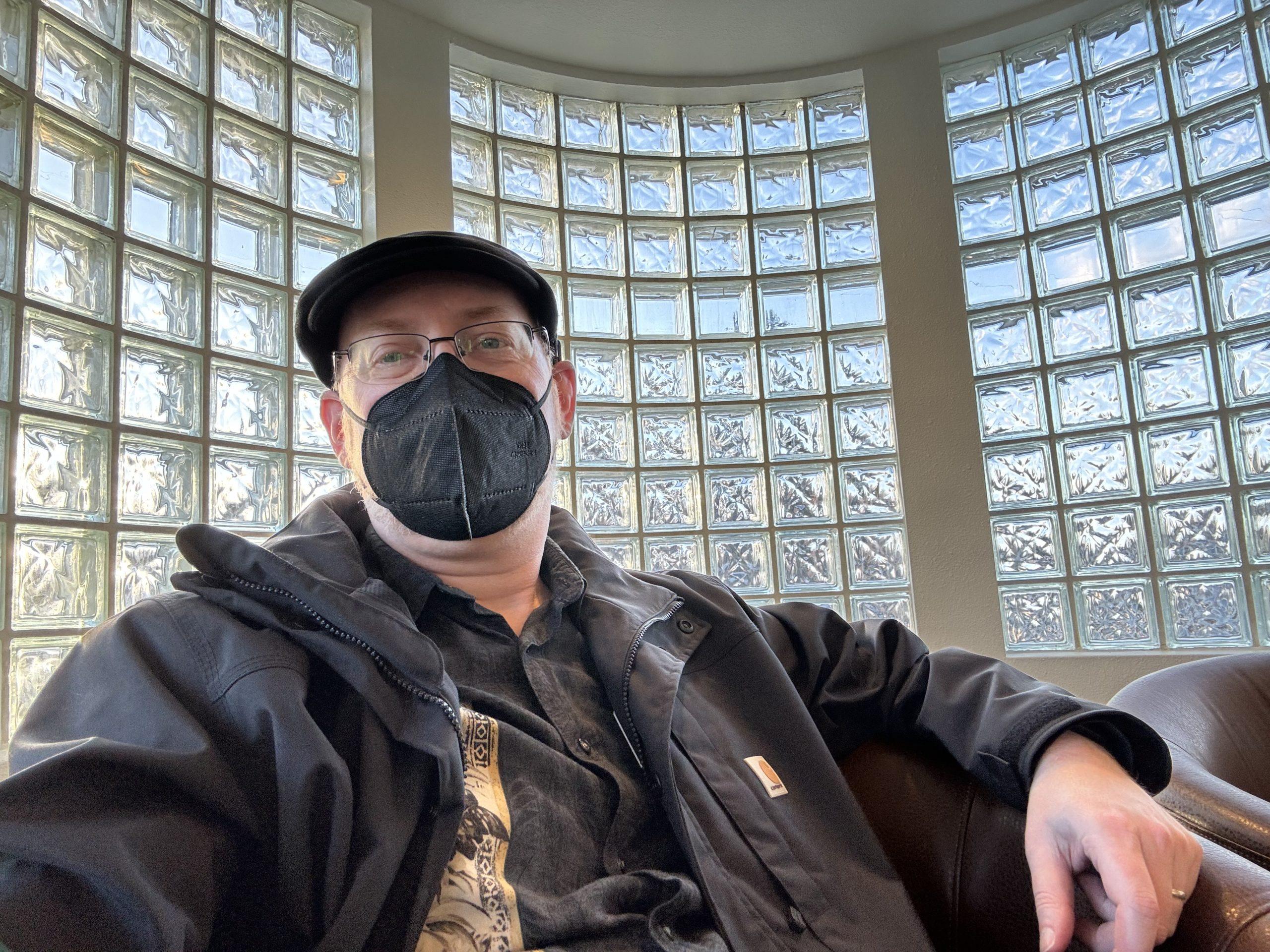 Me, wearing coat, hat, and face mask, sitting in front of a curved glass brick wall.
