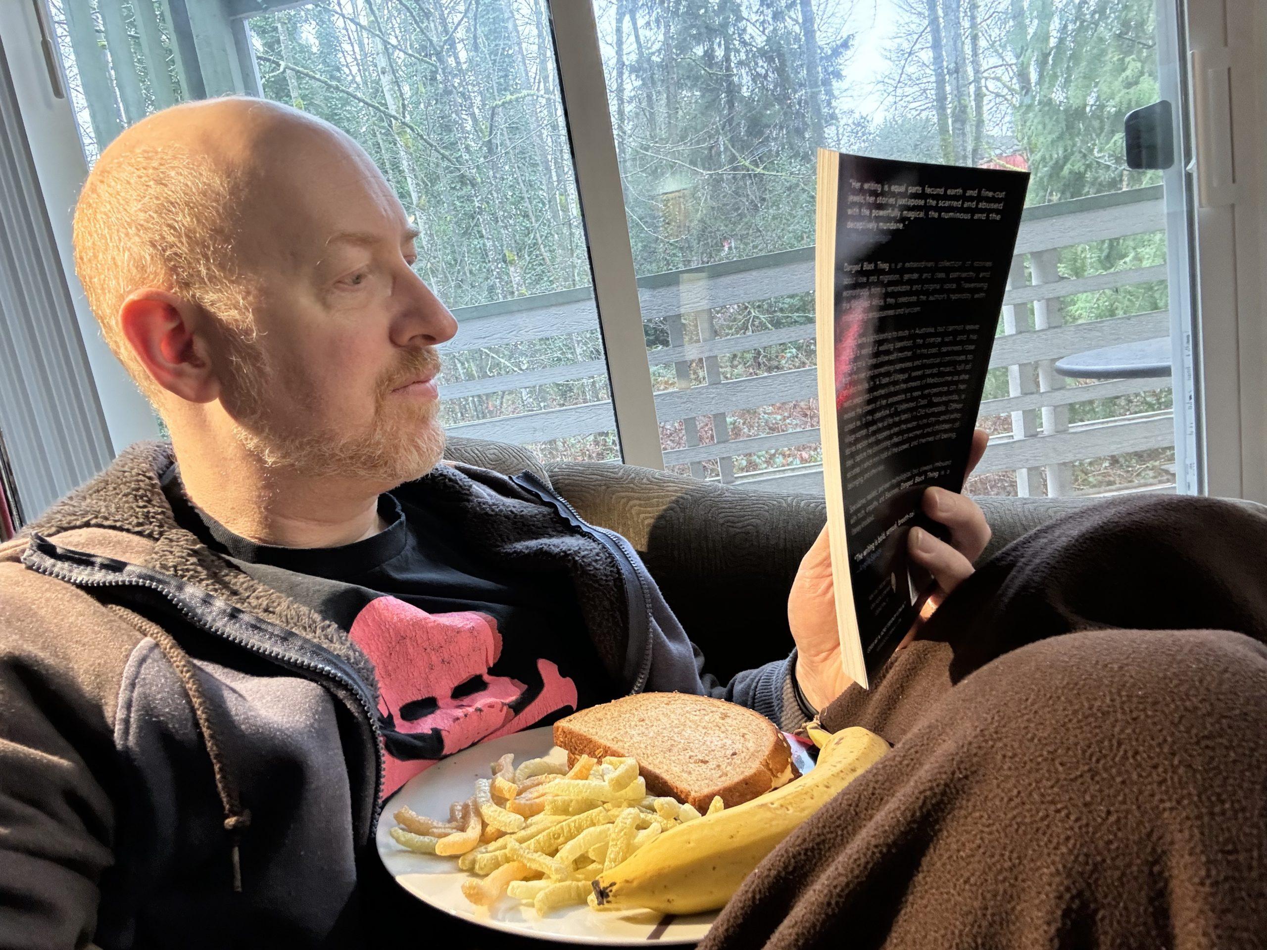 Me curled up in a chair with a lunch plate with sandwich, chips, and banana on my lap, reading a book.