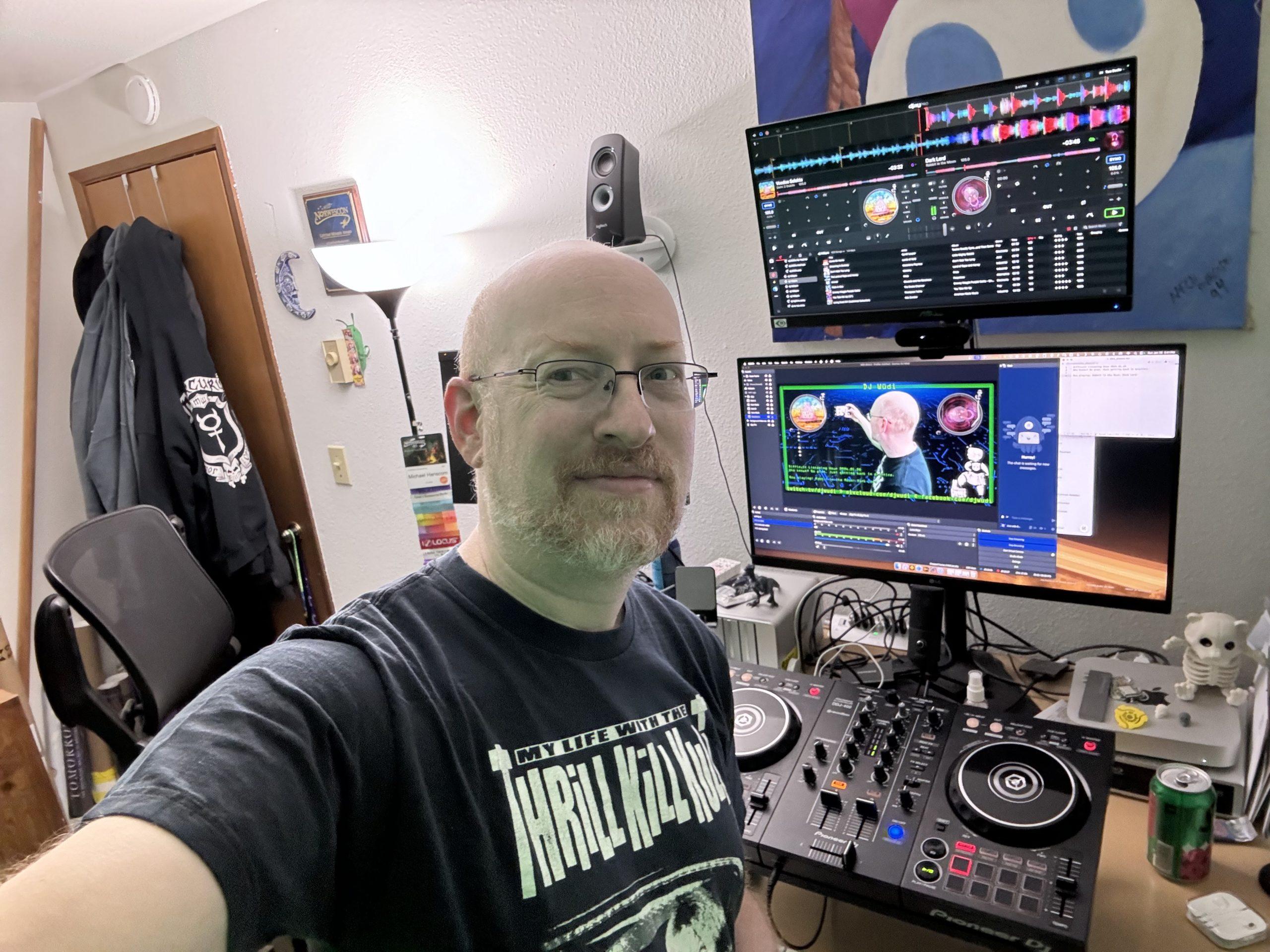 Me at my desk at home. Two monitors are mounted one above the other; the top monitor has DJ software, the bottom monitor has video broadcasting software. Below the monitors is a DJ controller.